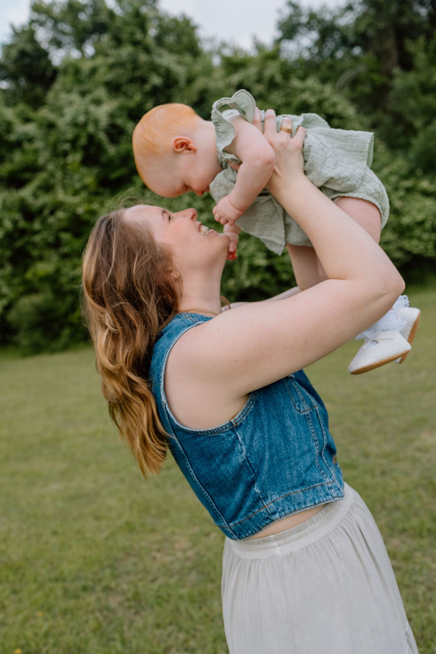 Person in denim vest lifts and kisses a baby wearing light colored clothing while standing in a green outdoor setting.