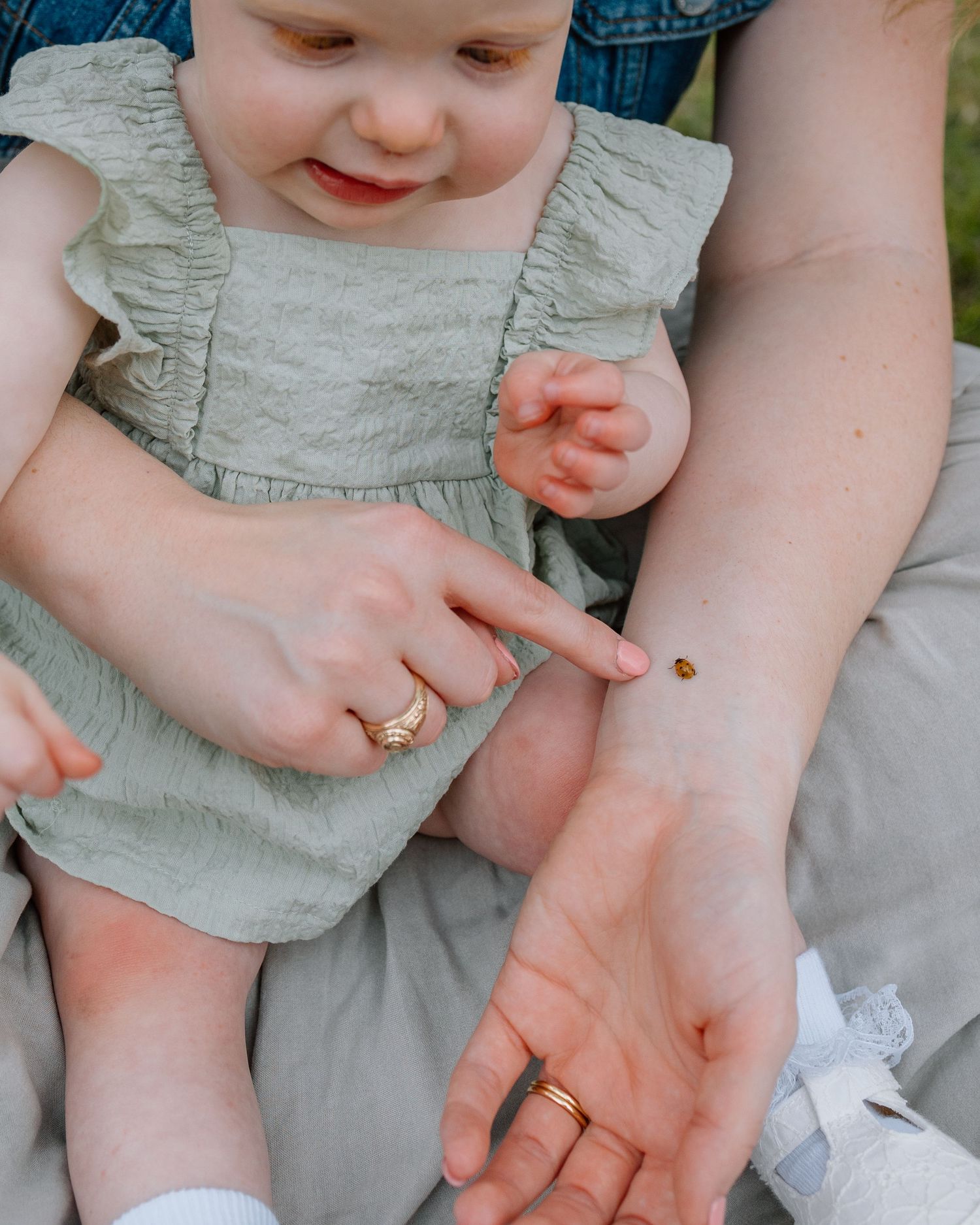 A curious child in a sage green romper points at a tiny ladybug resting on someone's arm.