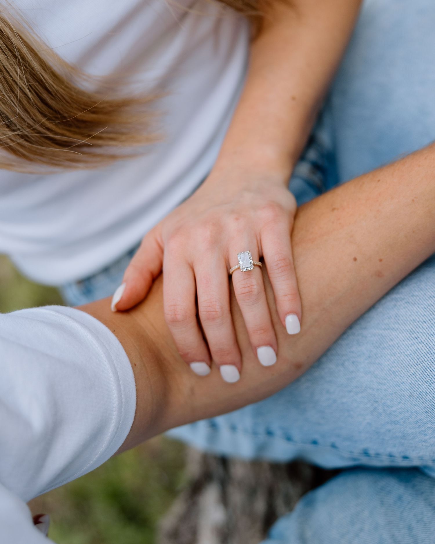 Close-up of a woman's hand displaying a gold diamond engagement ring while sitting outdoors in jeans.