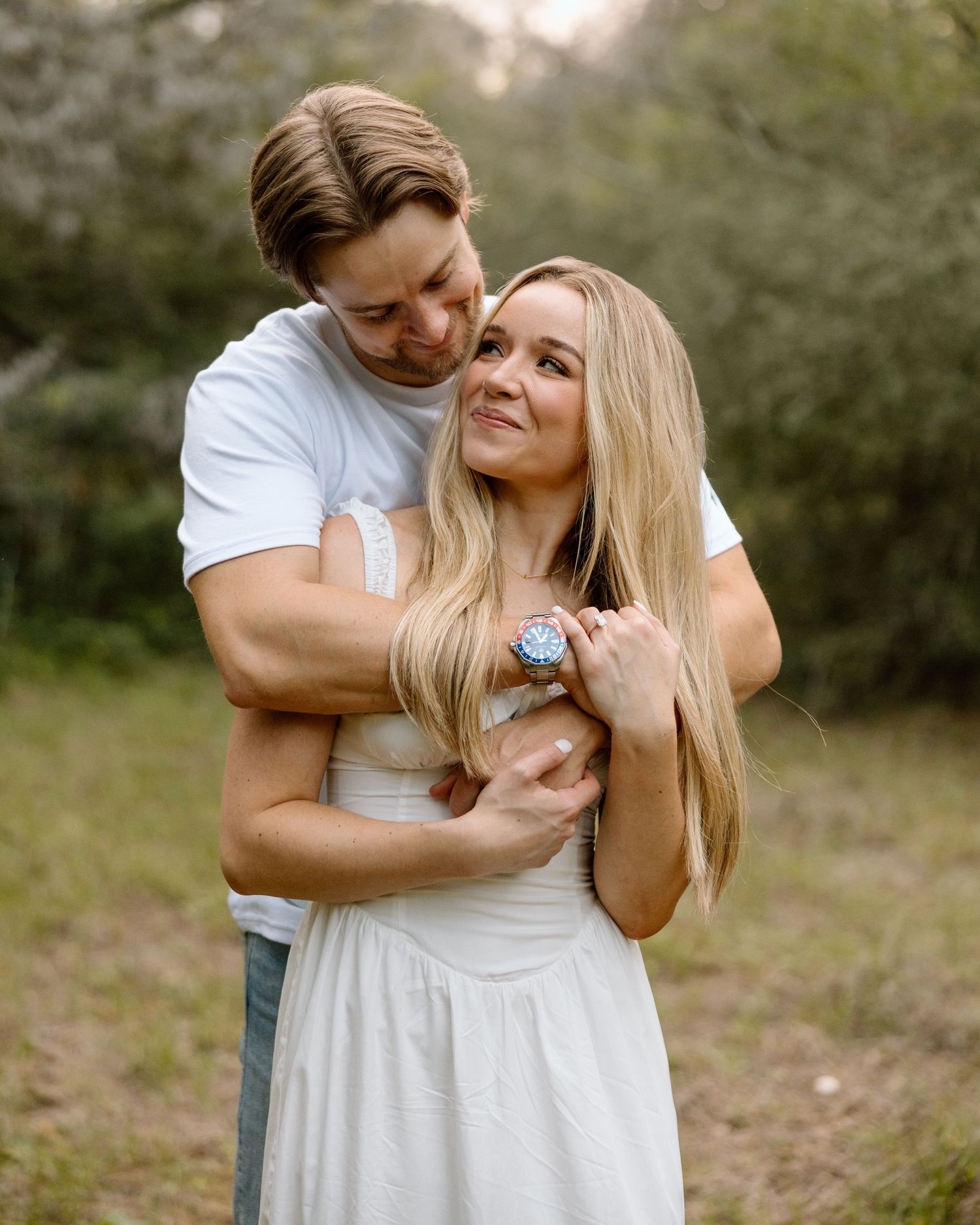 A couple embraces outdoors, man in white tee hugs blonde woman in white dress, surrounded by lush greenery.