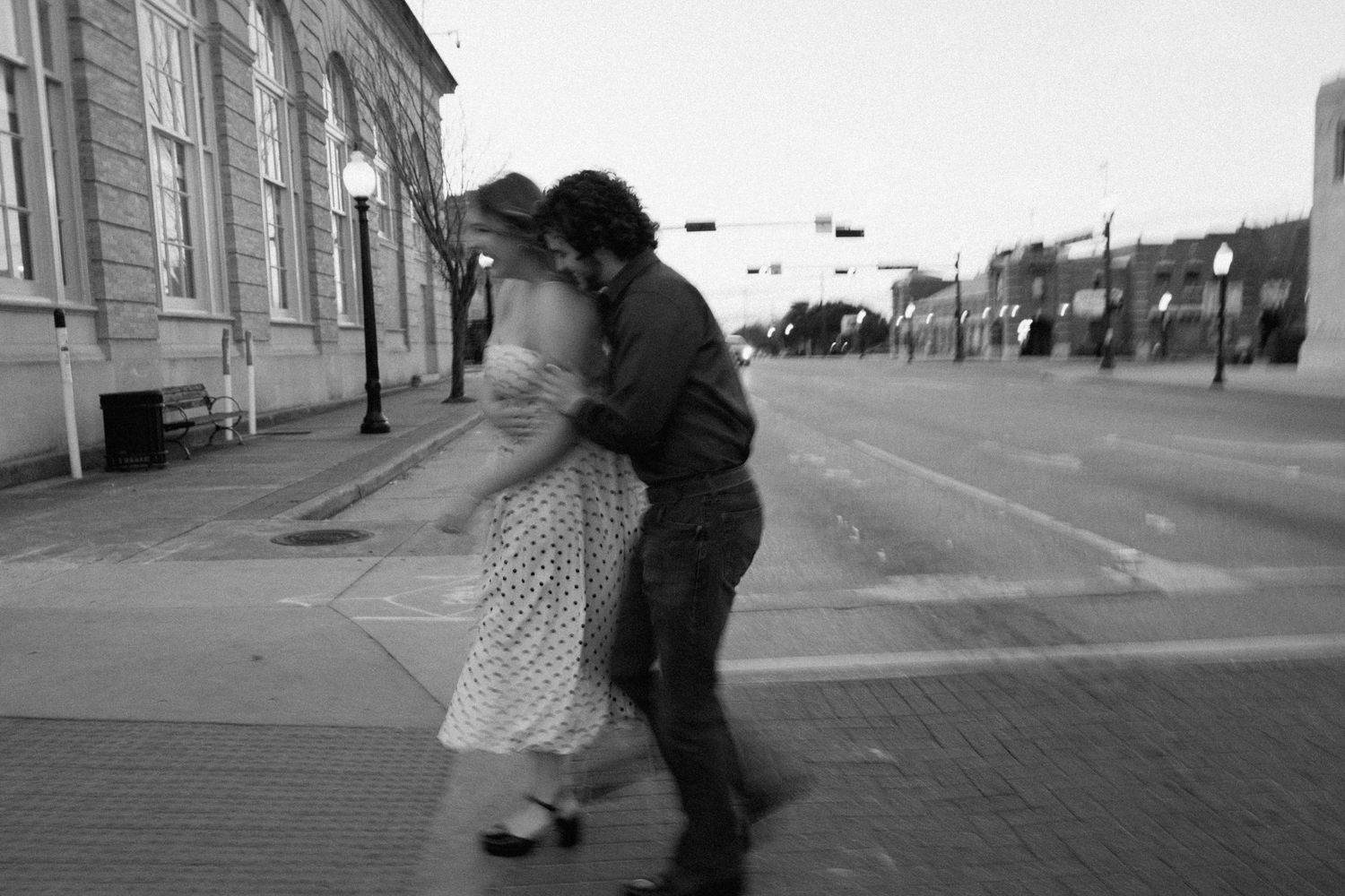Black and white photo of a couple dancing joyfully on a city street at dusk.