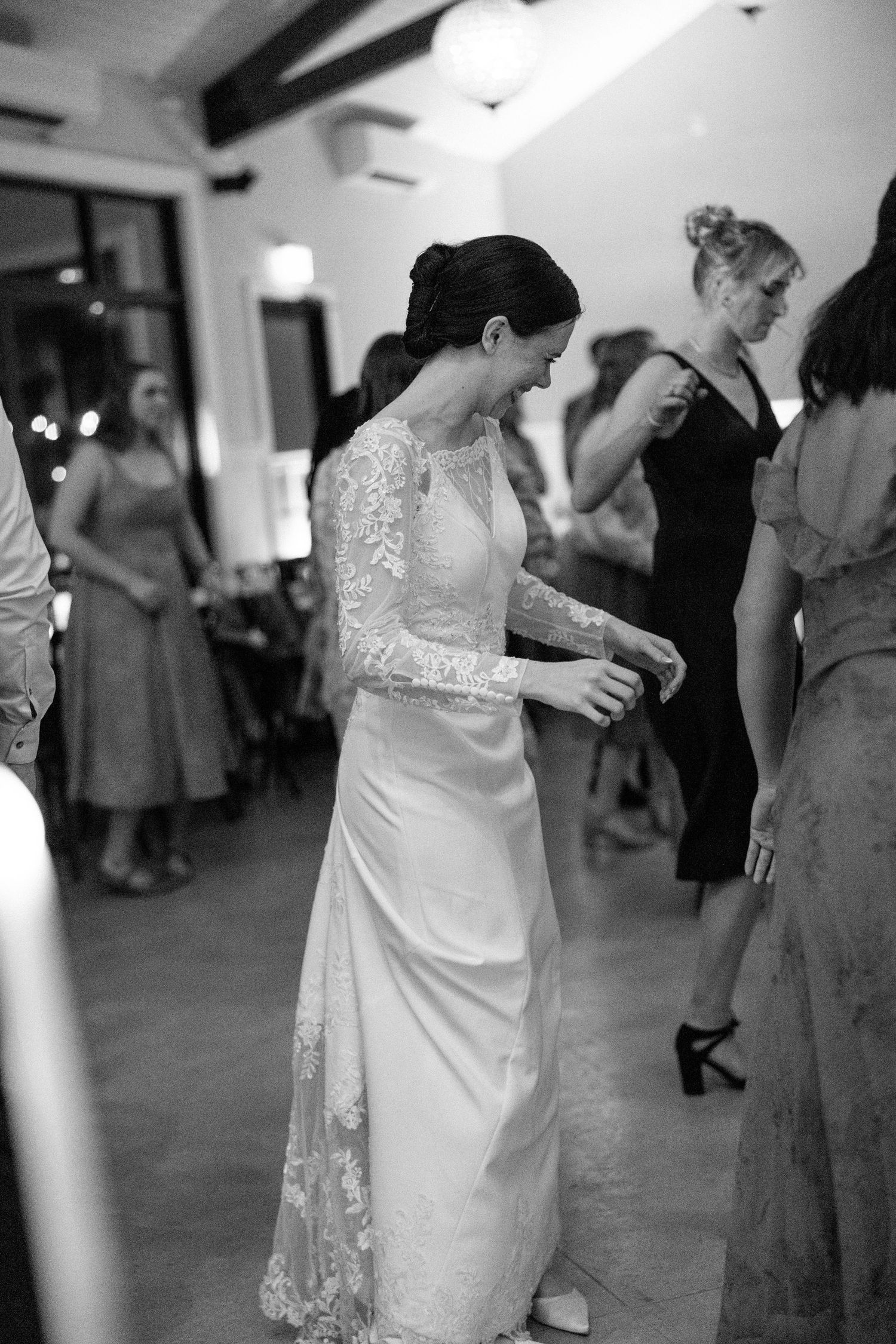 Bride in elegant lace wedding gown laughs and dances joyfully with guests at wedding reception.