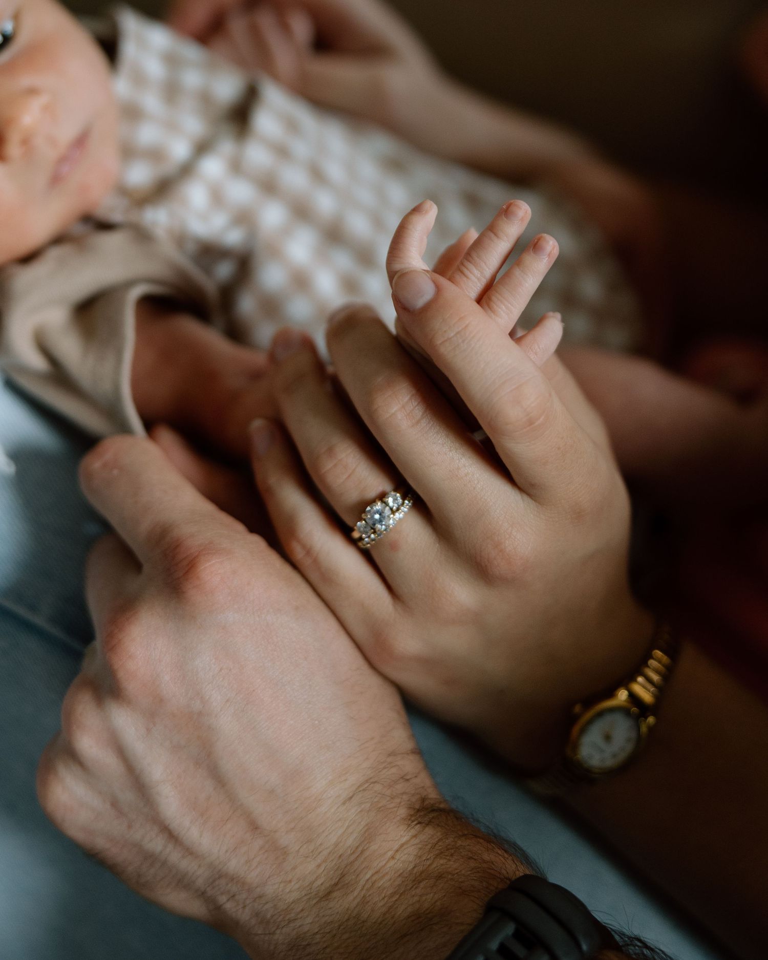 Close-up of parents' hands cradling newborn baby's tiny hand, mother wearing diamond ring and gold watch.