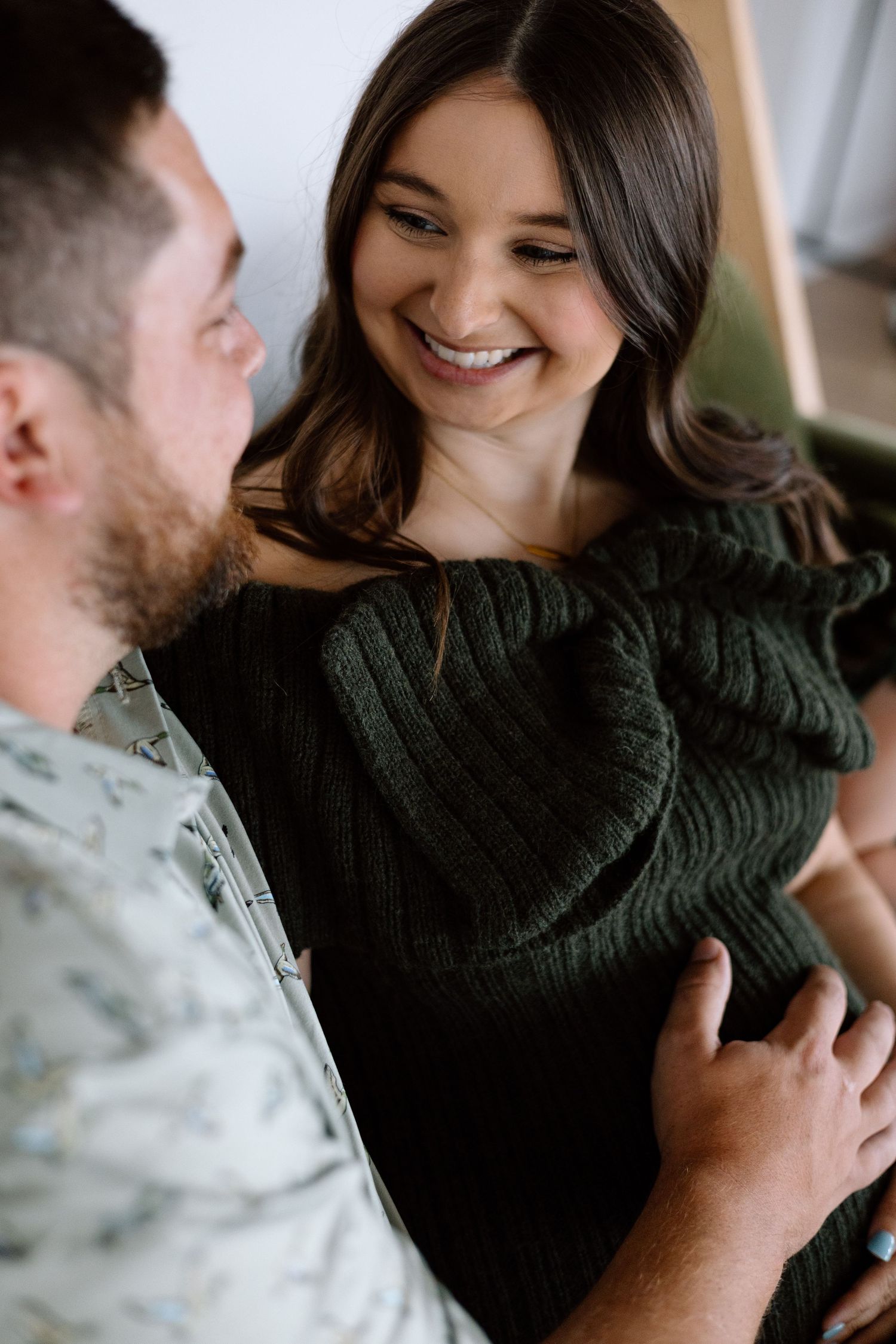 A smiling woman in a dark green knit sweater shares a tender moment with a bearded man touching her baby bump.