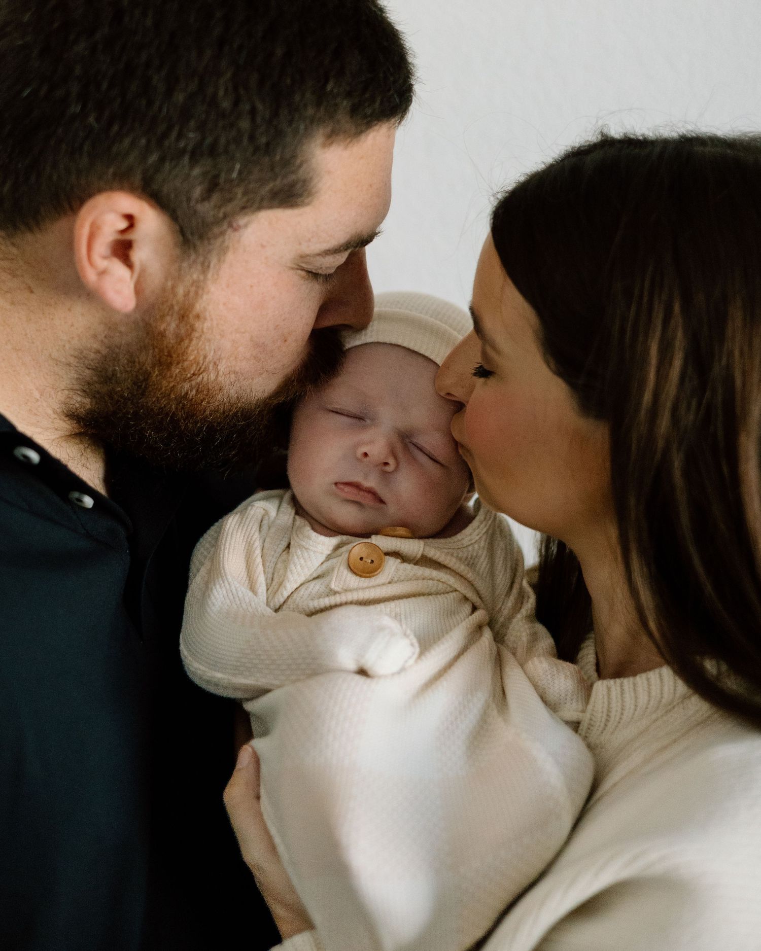 New parents tenderly kissing their sleeping newborn dressed in cream knit outfit.
