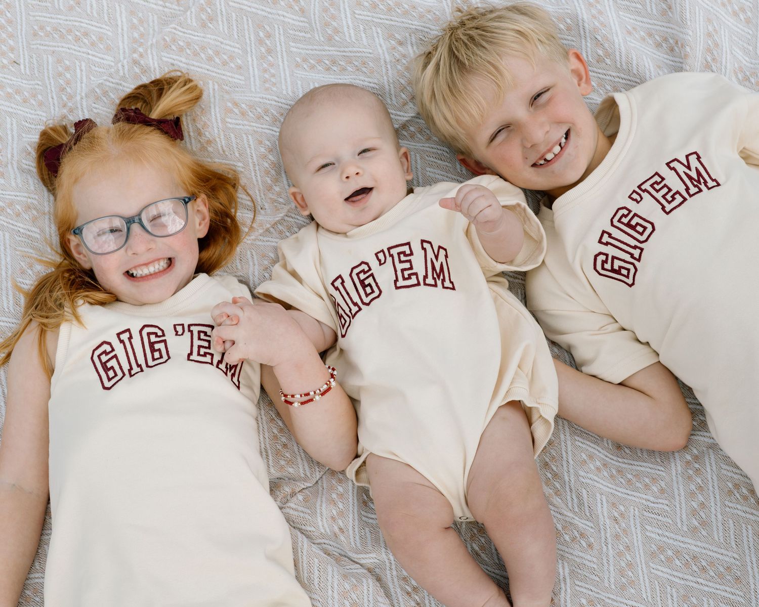Three children wearing matching cream GIG'EM shirts lying together, smiling happily in a cute sibling photo.