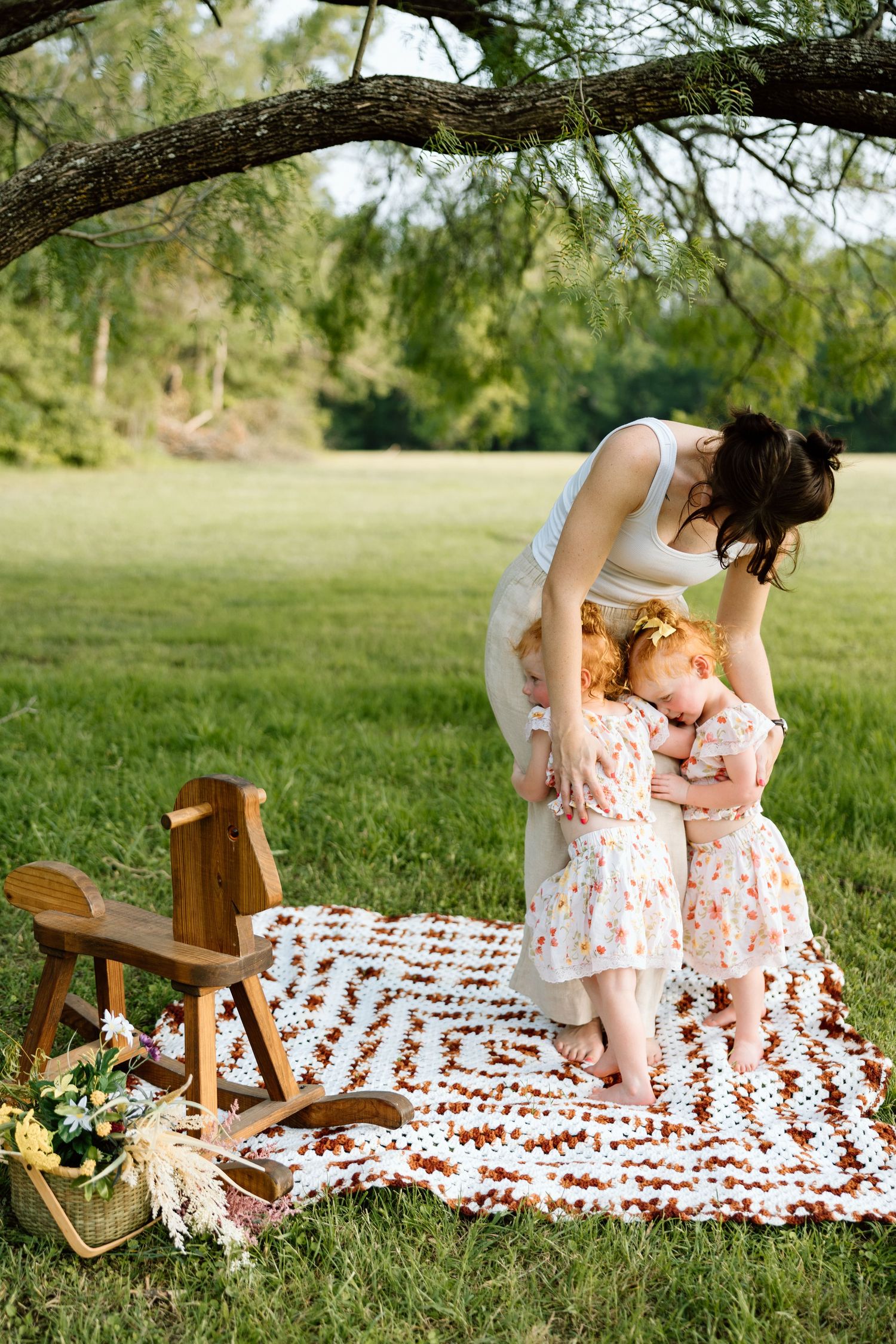 Mother and two young girls in floral dresses sharing a tender moment on a patchwork blanket outdoors.