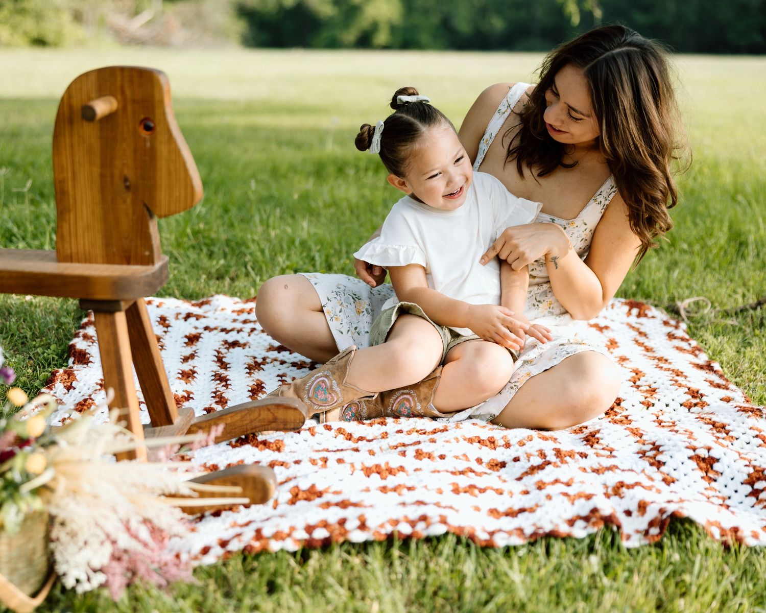 A mother and young child share a joyful moment on a crochet blanket during an outdoor picnic on green grass.