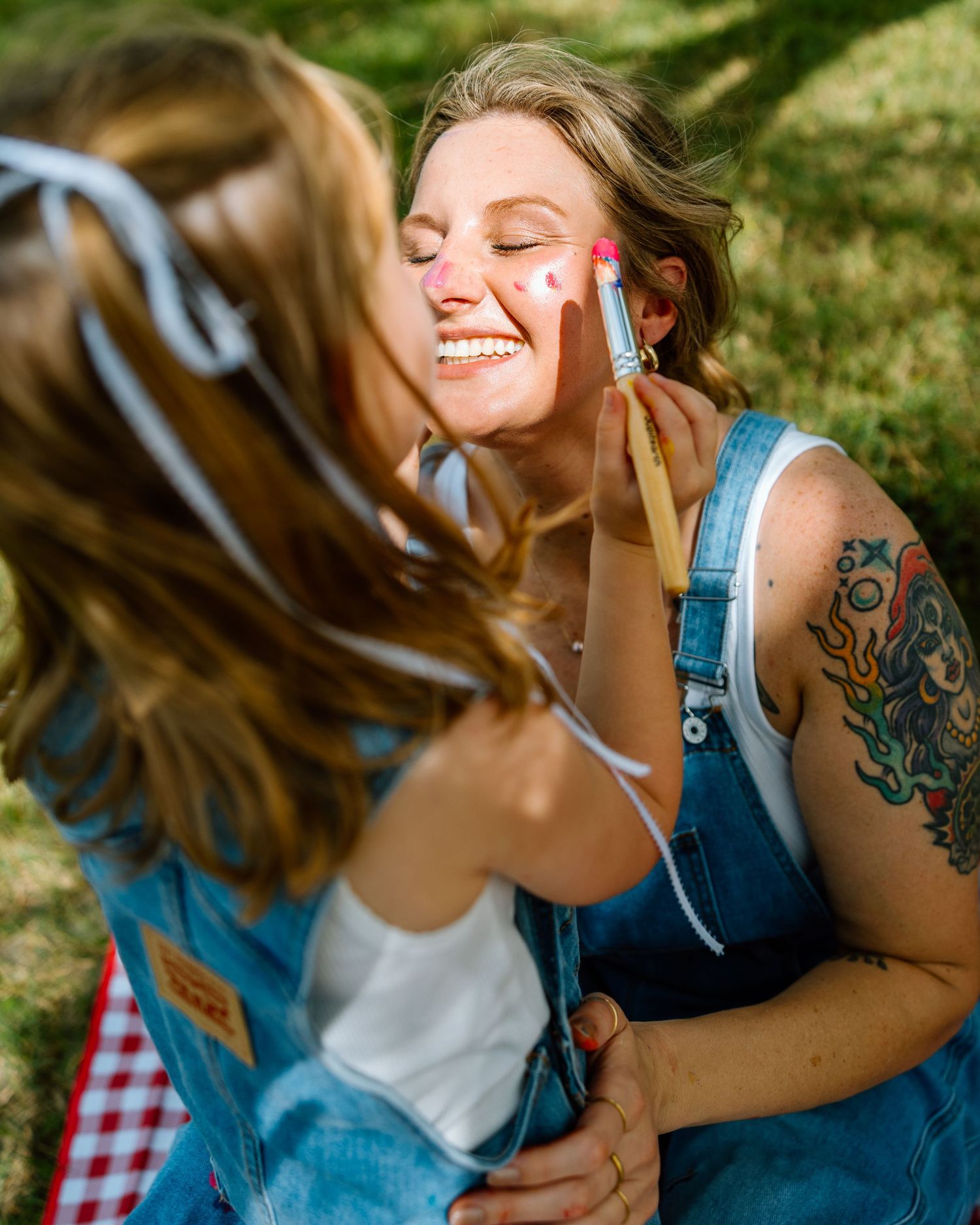 A smiling woman in denim overalls with a colorful tattoo gets her face painted outdoors on a sunny day.