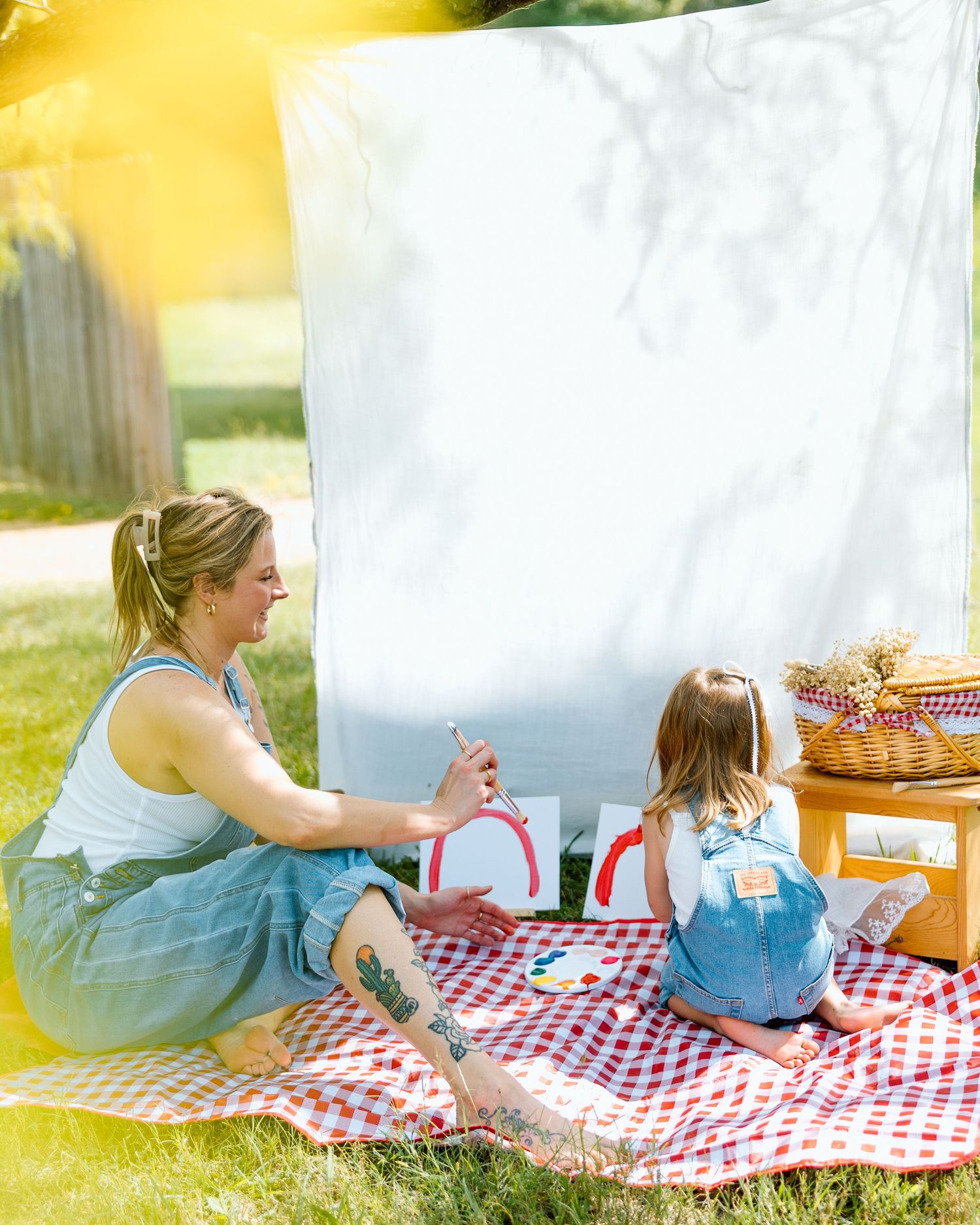 Woman and young girl painting together on a red gingham picnic blanket outdoors on a sunny day.