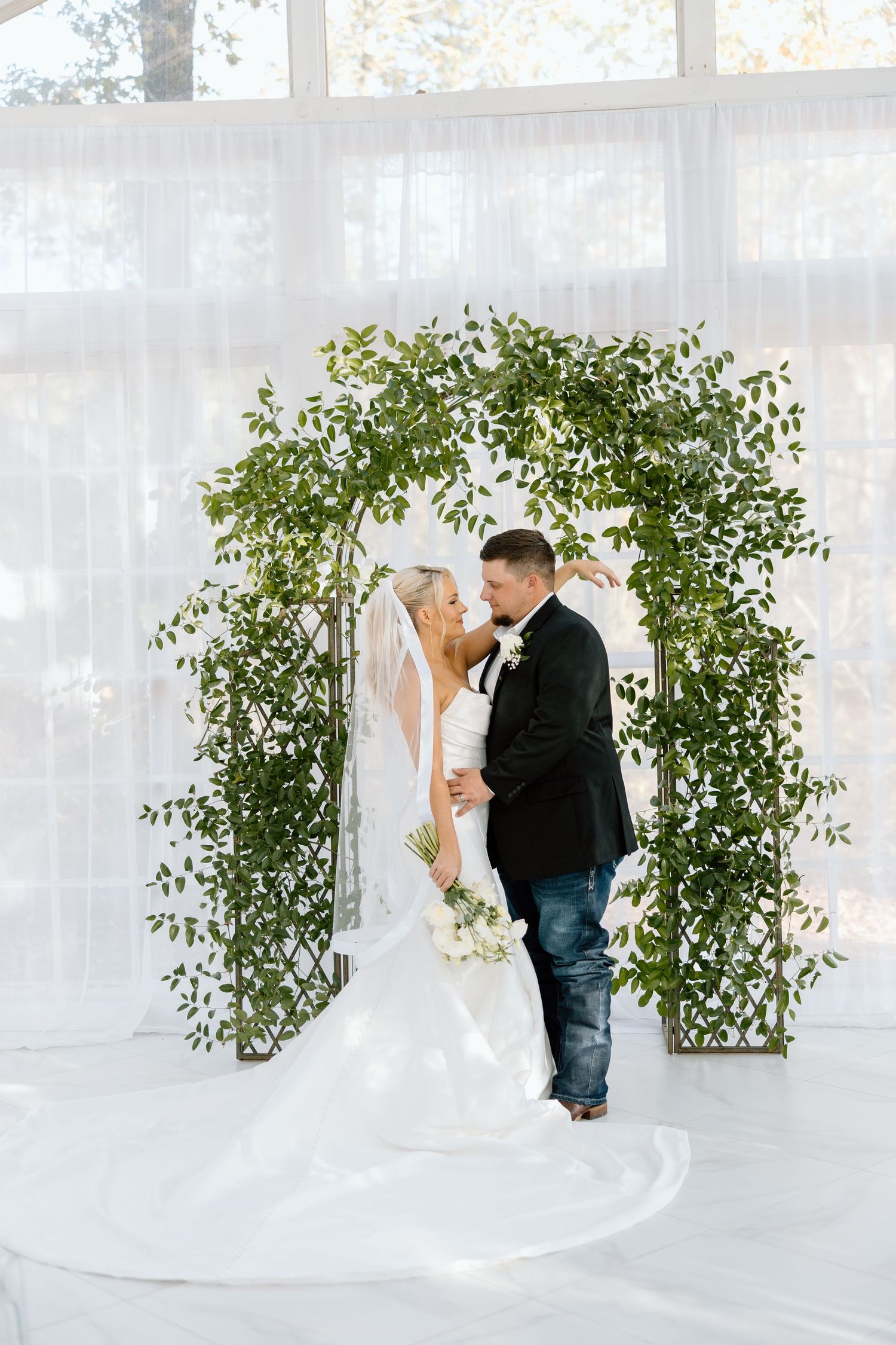Bride and groom share a romantic moment under a greenery arch with white draping at their elegant wedding ceremony.