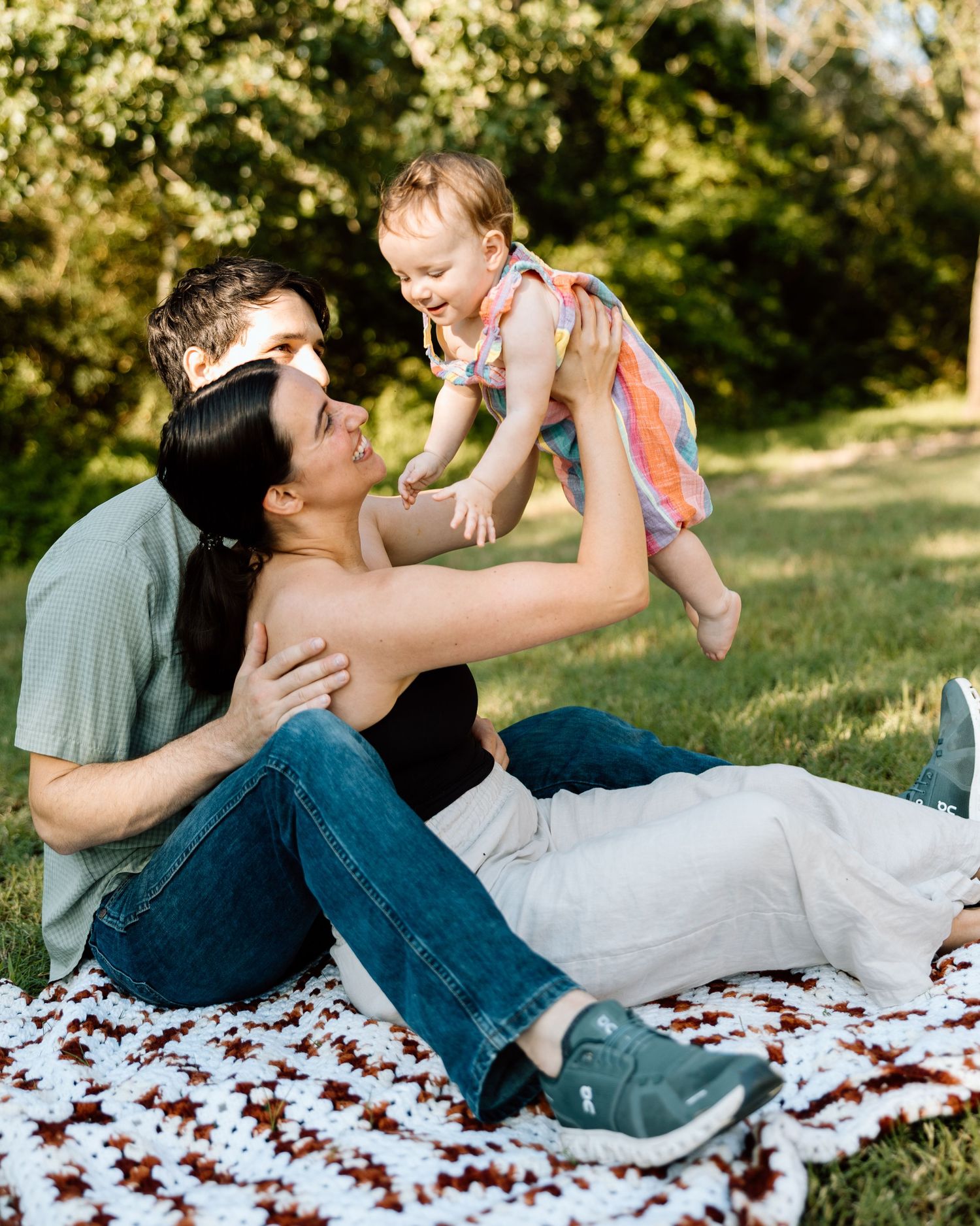 A happy young family sits on a blanket in a sunny park, with parents lifting their smiling baby girl in a colorful dress.