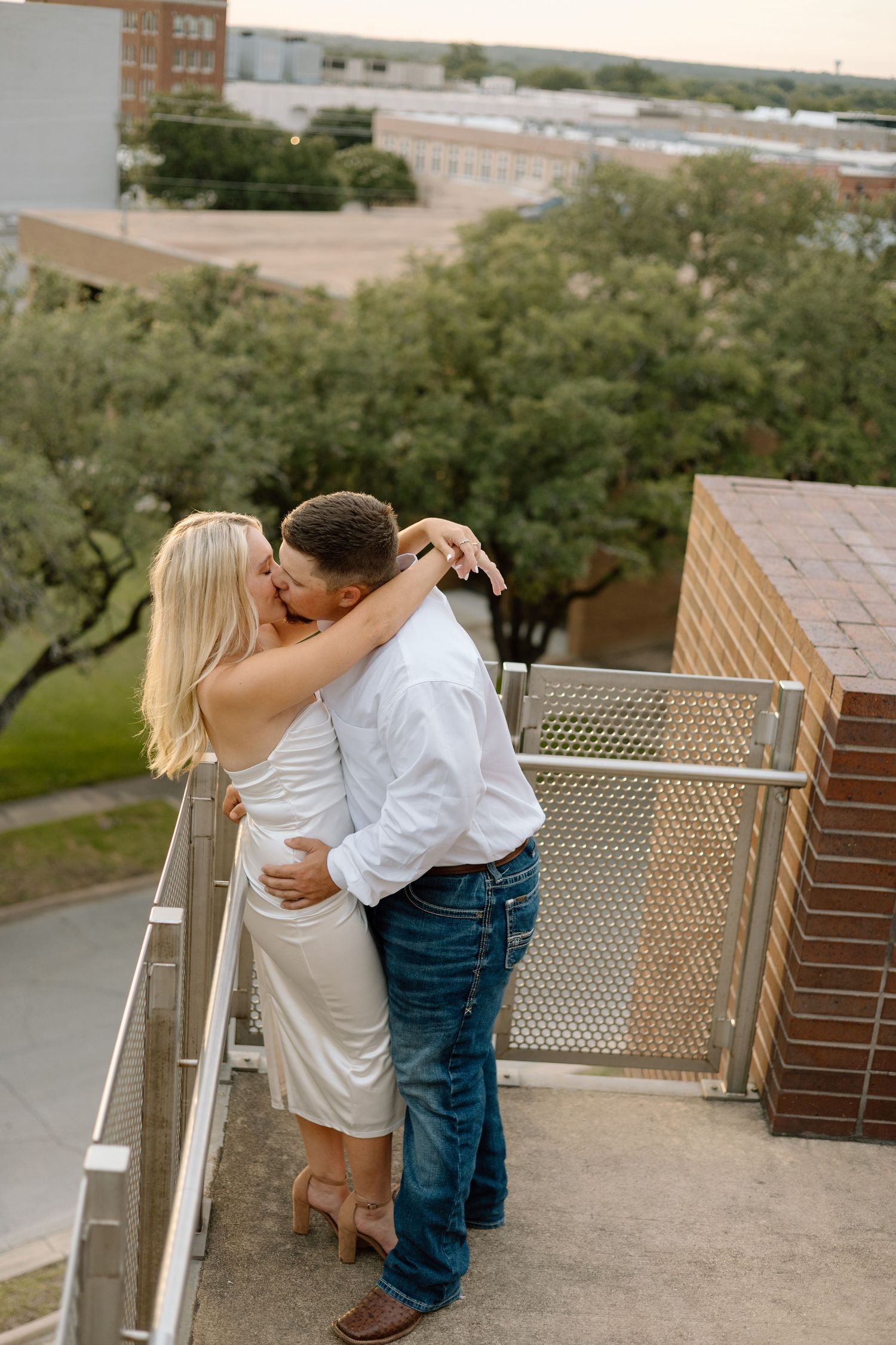 Couple sharing a romantic kiss on a rooftop balcony, woman in white dress and man in white shirt and jeans.