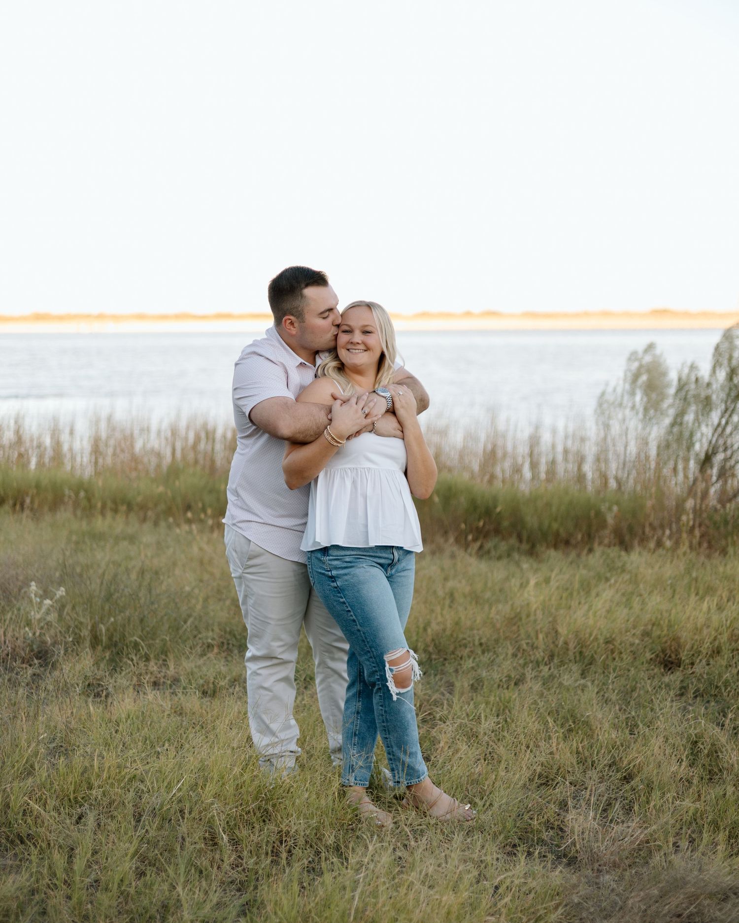 Couple embracing in a grassy field near a lake, man kissing woman's cheek during golden hour engagement photo.