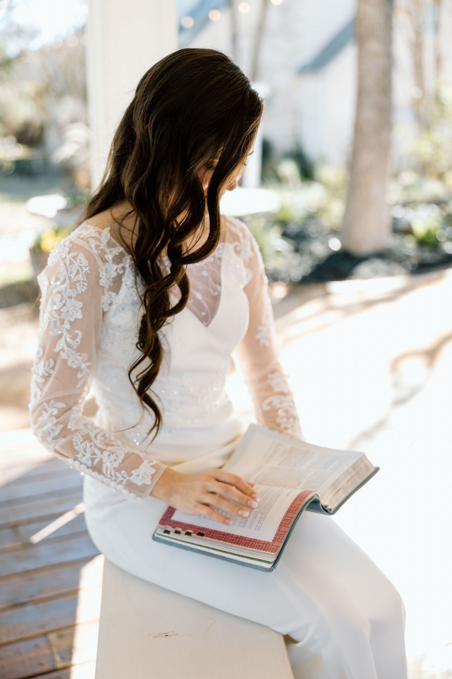 Bride in lace long-sleeve wedding gown reading a book outdoors before her ceremony.