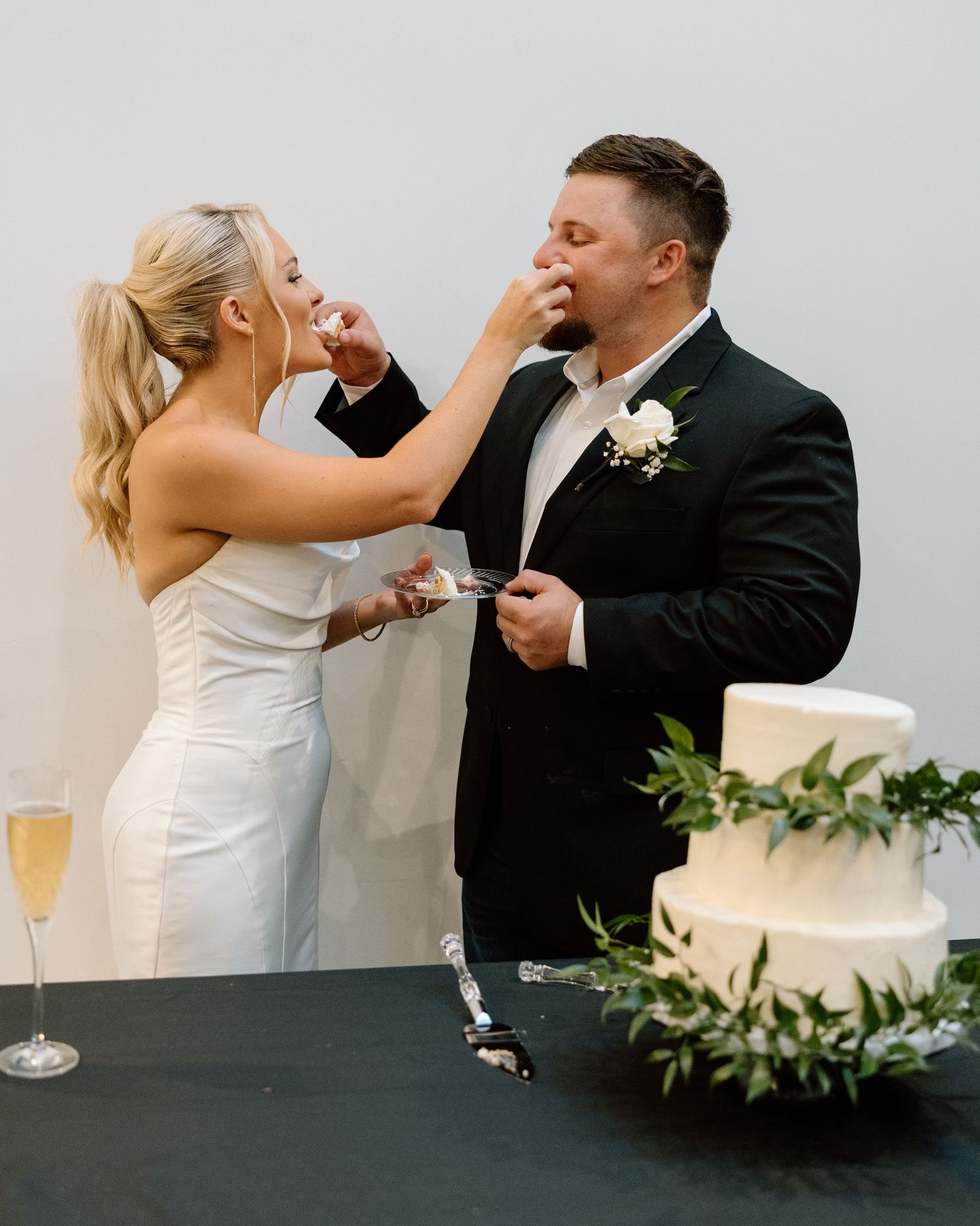 Blonde bride feeds wedding cake to groom beside a white three-tier cake with greenery decor.
