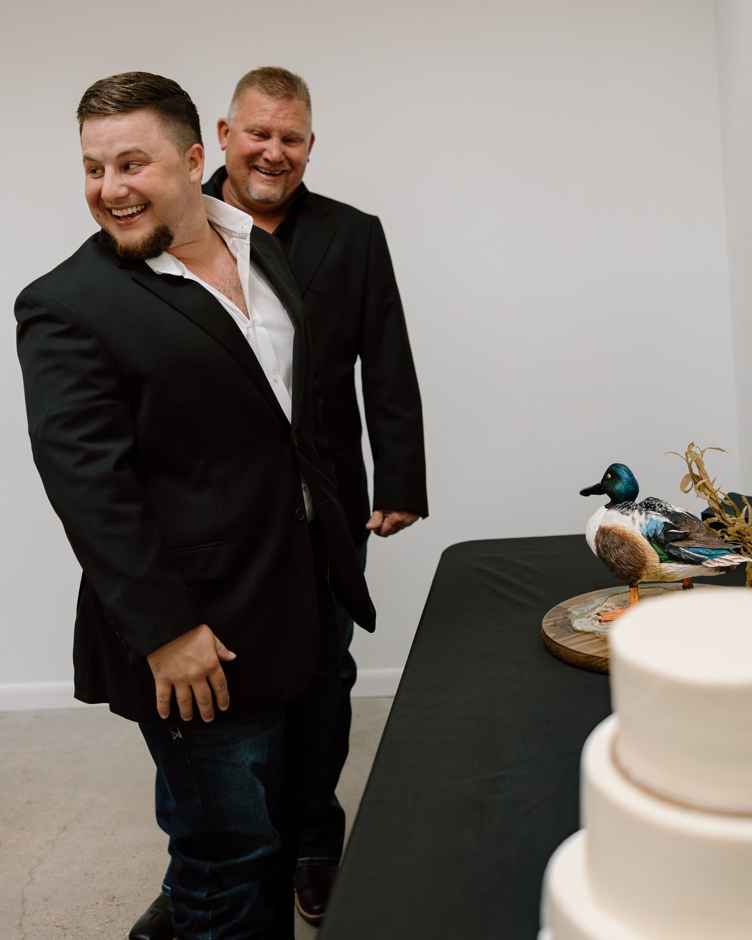 Two men in black suits smiling near a wedding cake with a duck decoy topper at a reception.