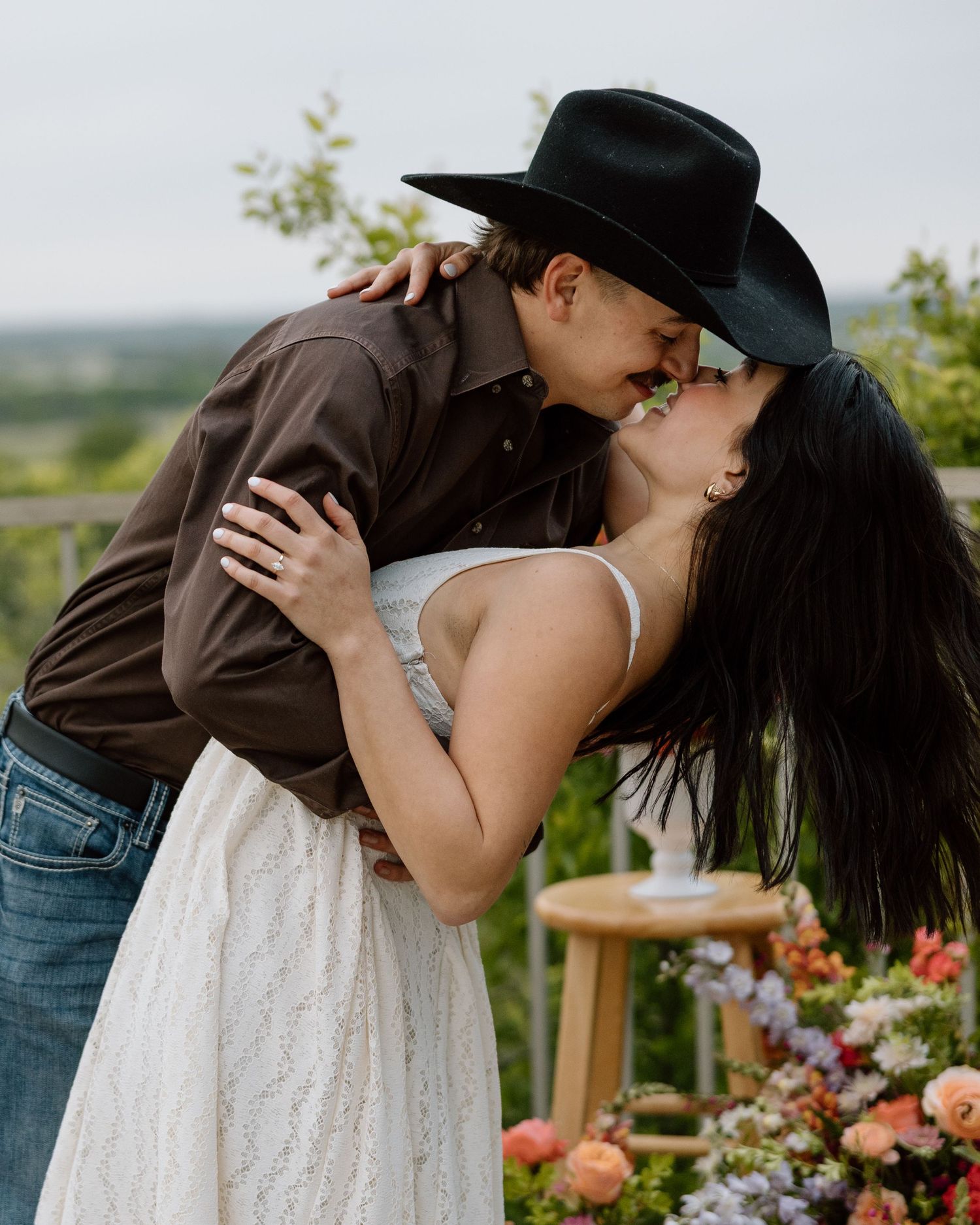 Couple sharing a romantic kiss outdoors, man in black cowboy hat and brown shirt, woman in white lace dress, floral arrangement nearby.