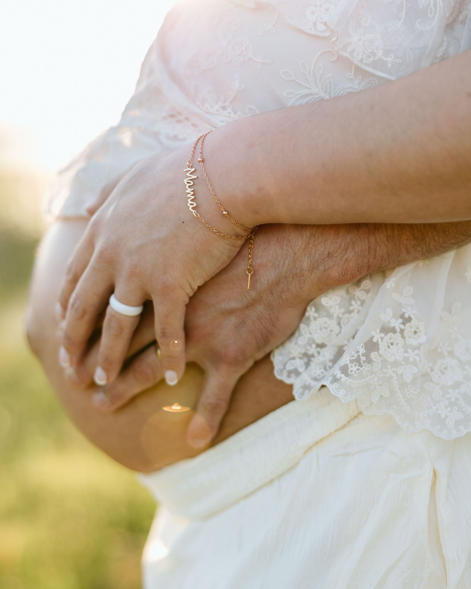 Pregnant woman in white lace dress with gold mama bracelet, partner's hand on baby bump, outdoor maternity photo.