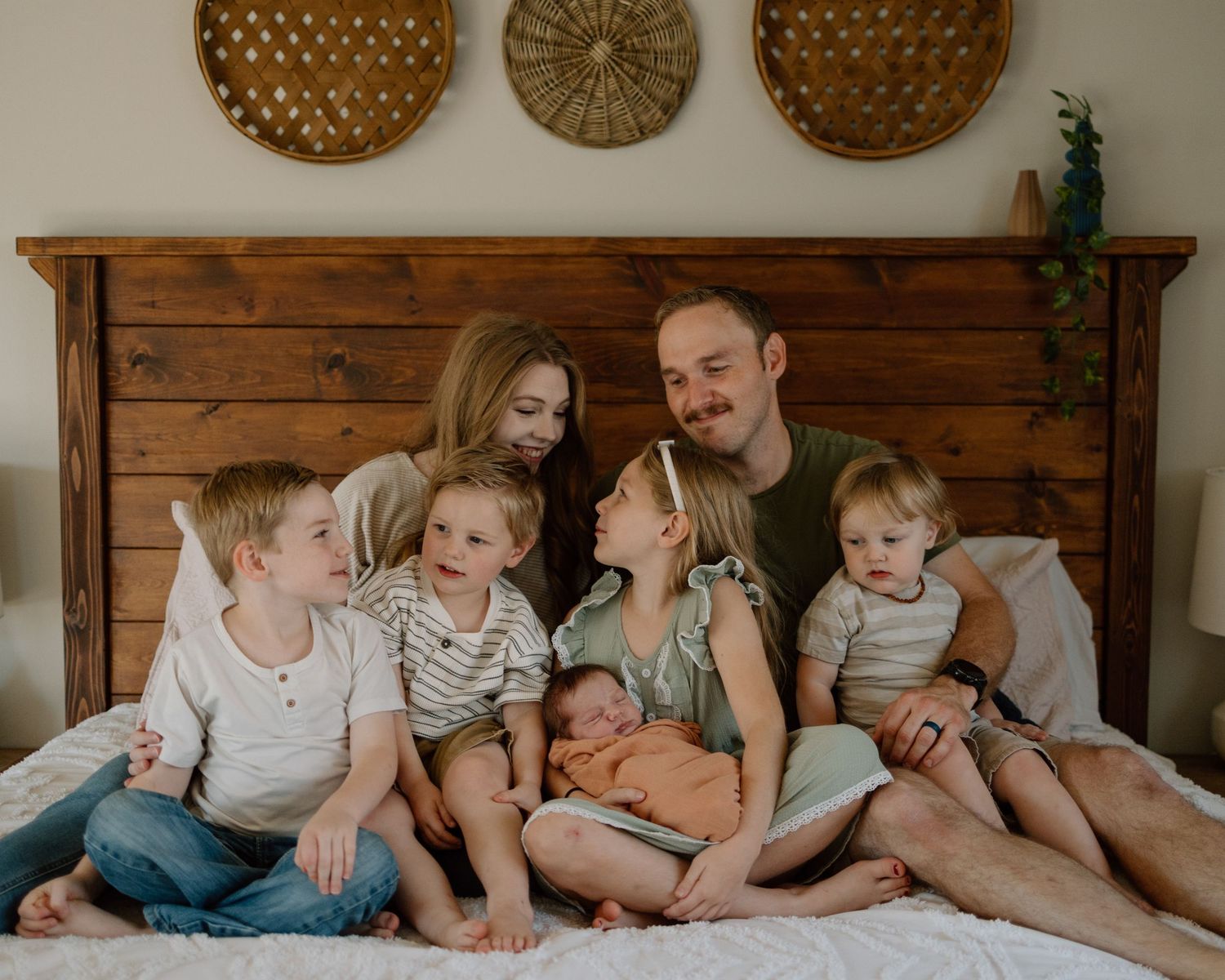 A cozy family photo session on a bed with rustic wooden headboard and woven baskets displayed on the wall above.