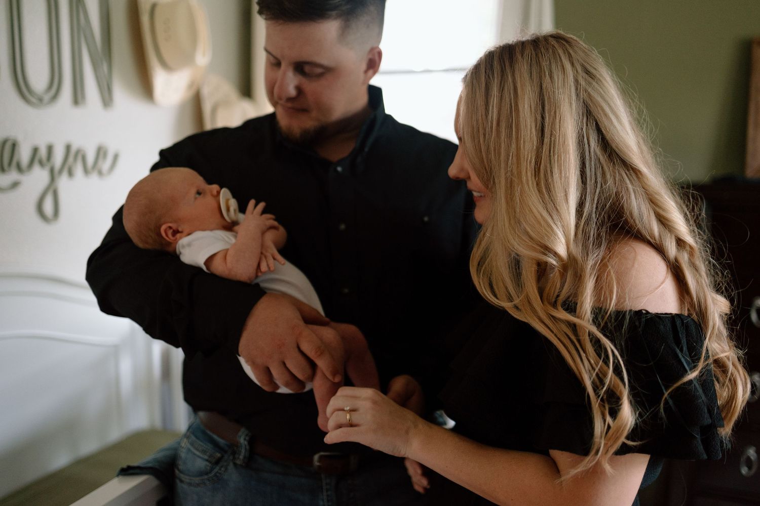 A couple in black outfits tenderly cares for their newborn baby in a softly lit room.