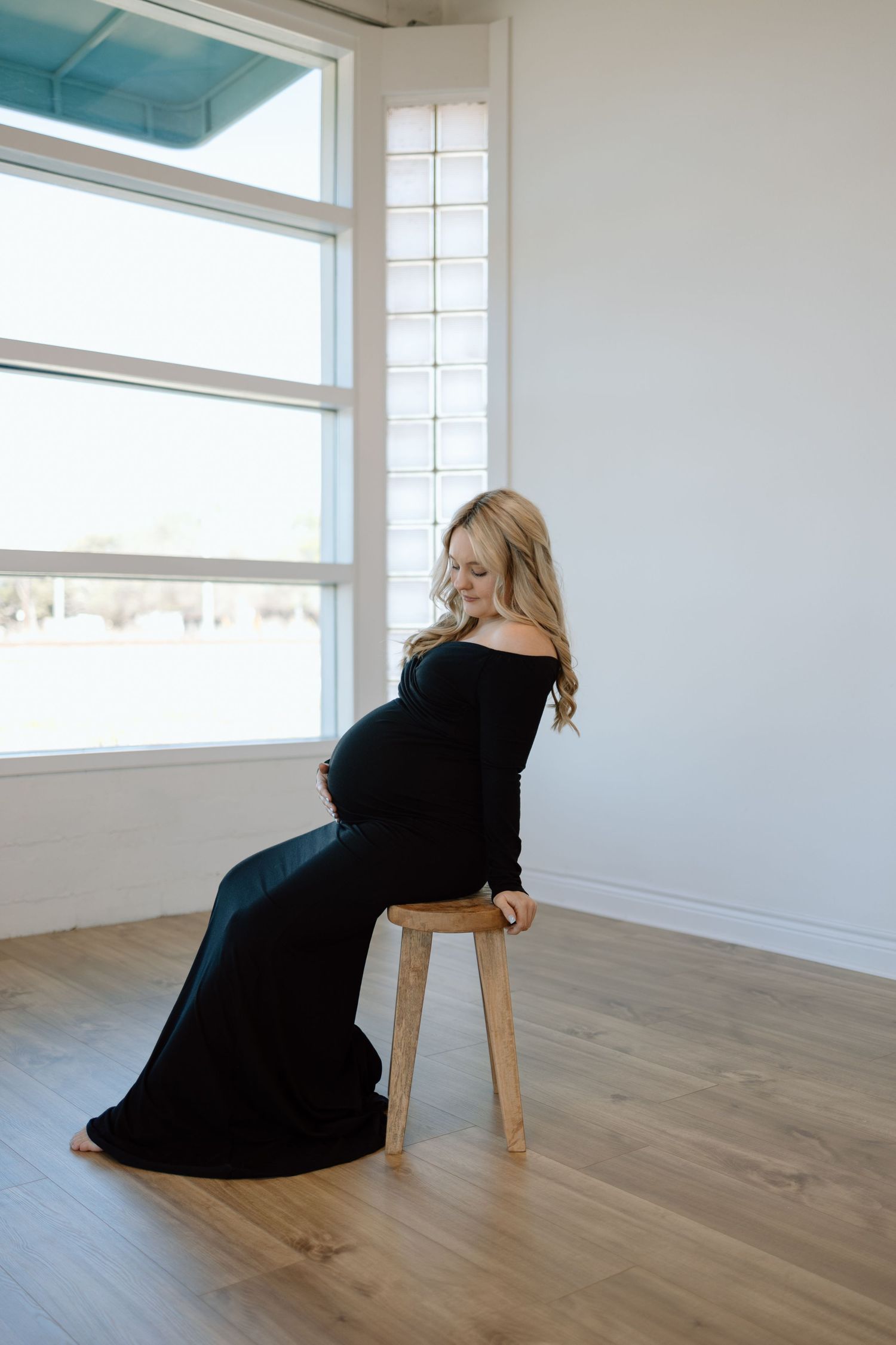 Maternity photo showing expectant mother in elegant black off-shoulder dress seated on wooden stool by bright windows.