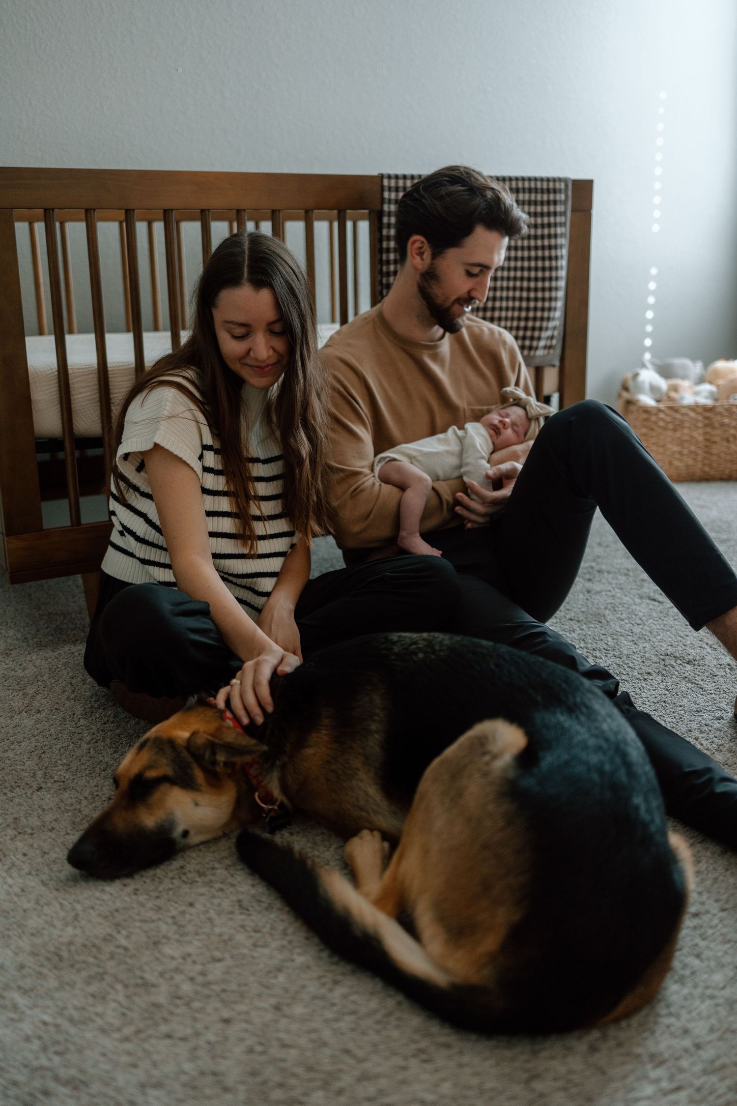 Family relaxing on floor with their German Shepherd dog while holding newborn baby near wooden crib.