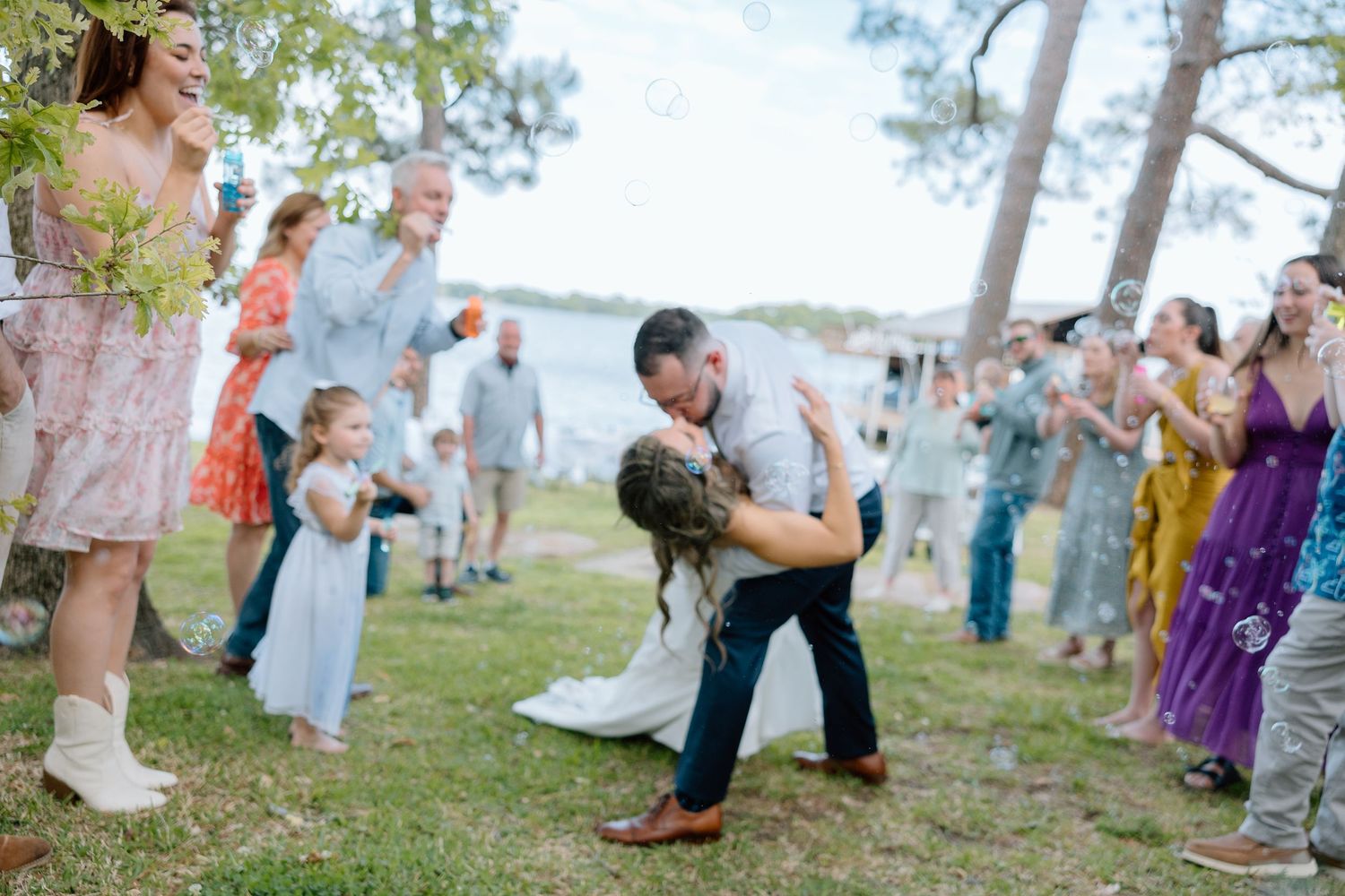 Wedding guests celebrating and dancing outdoors near pine trees and water.