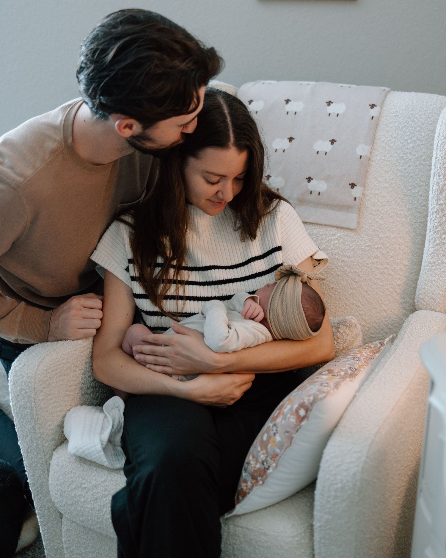 Family cuddles together on white armchair while holding their newborn.
