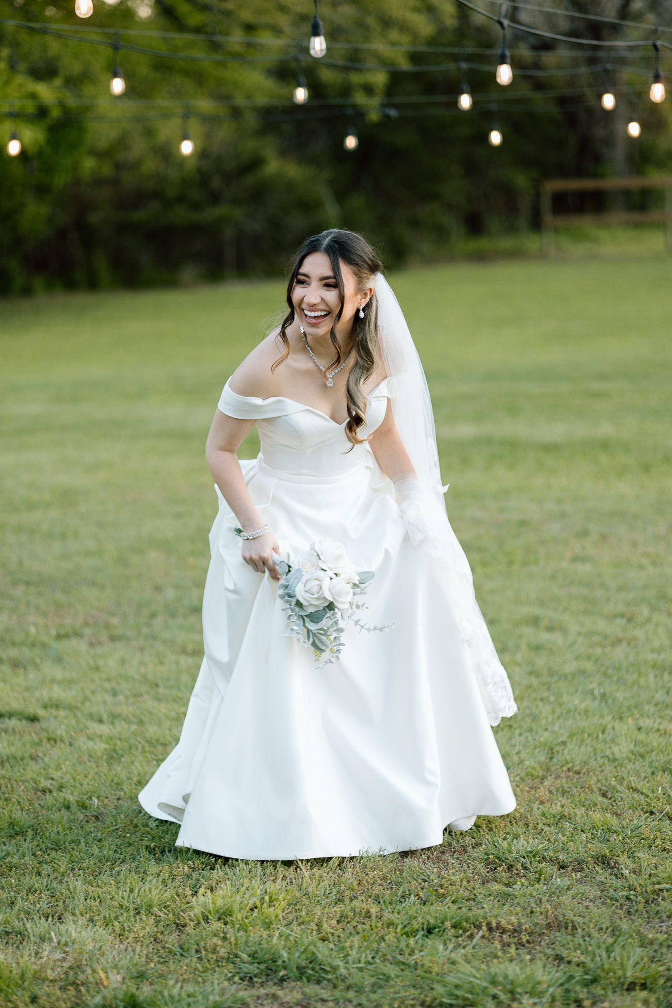 Bride in off-shoulder wedding gown laughs while holding bouquet under string lights.