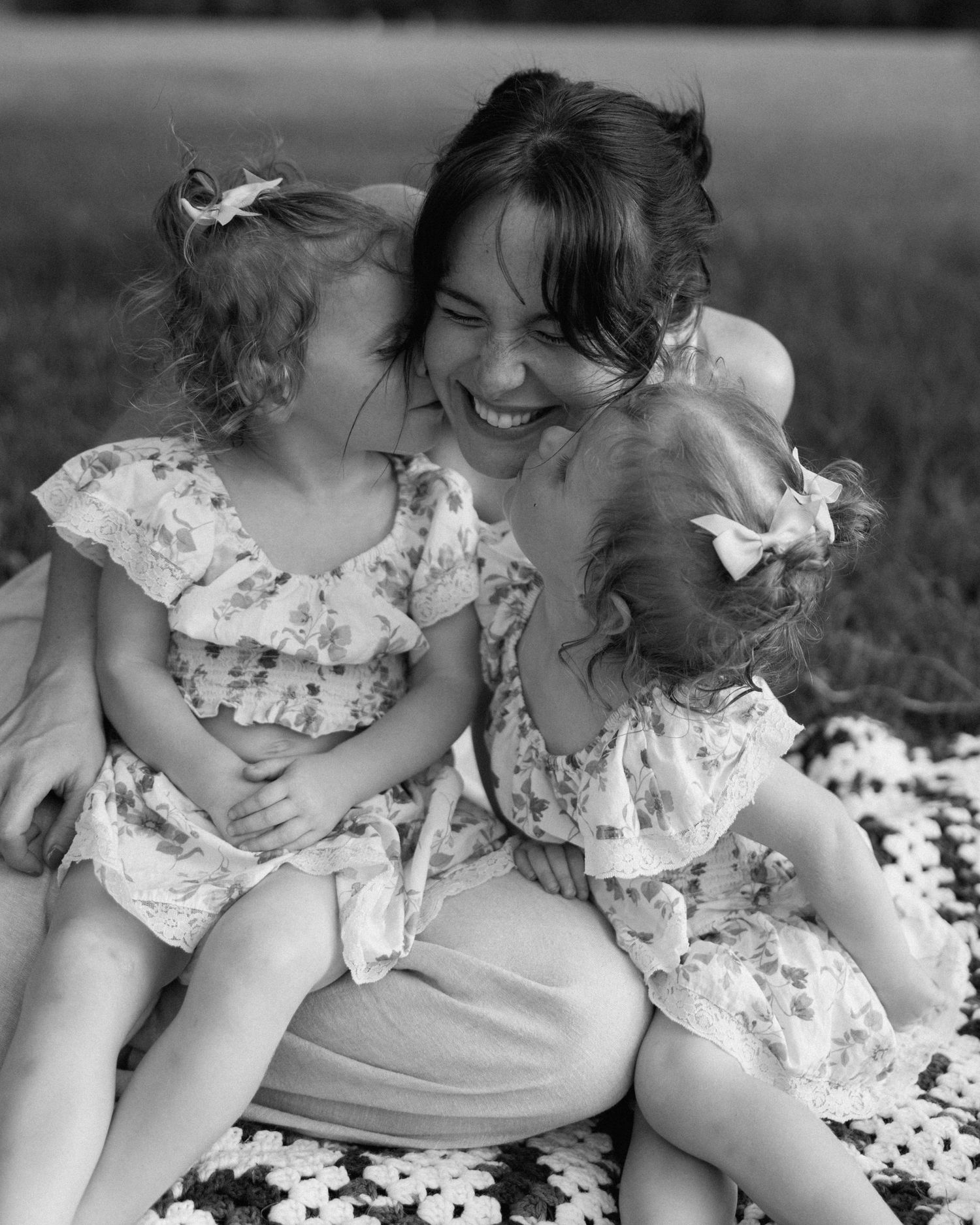 Black and white photo of a laughing mother with two young girls in matching floral dresses sitting outdoors.