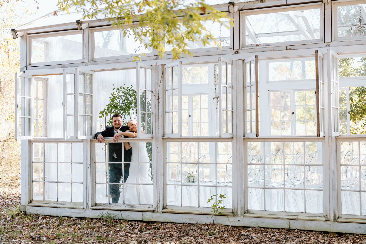 Bride and groom share a romantic moment inside a rustic white greenhouse with large vintage windows.