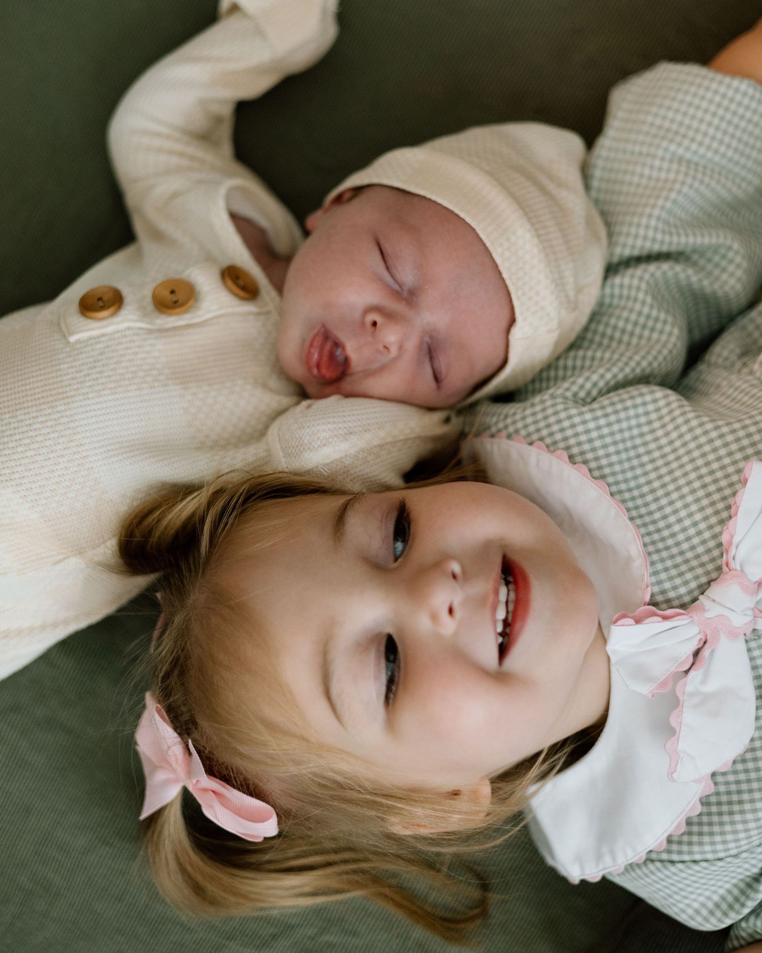 Smiling toddler girl with pink bow lying next to sleeping newborn baby sibling.