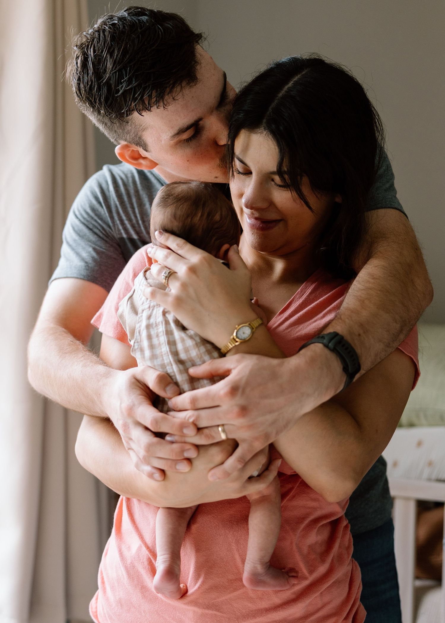 Young couple sharing a tender moment, kissing while lovingly holding their newborn baby together indoors.