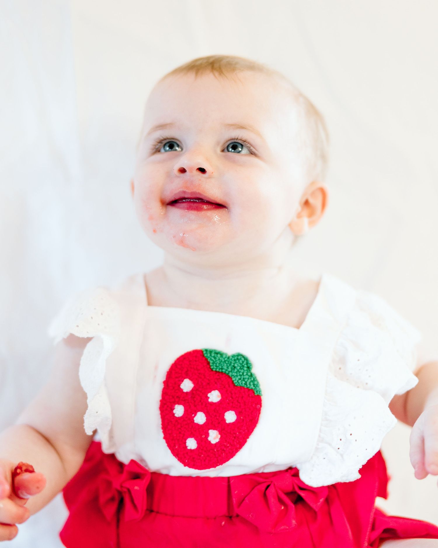Smiling baby in white strawberry applique top and red skirt looking up cheerfully.