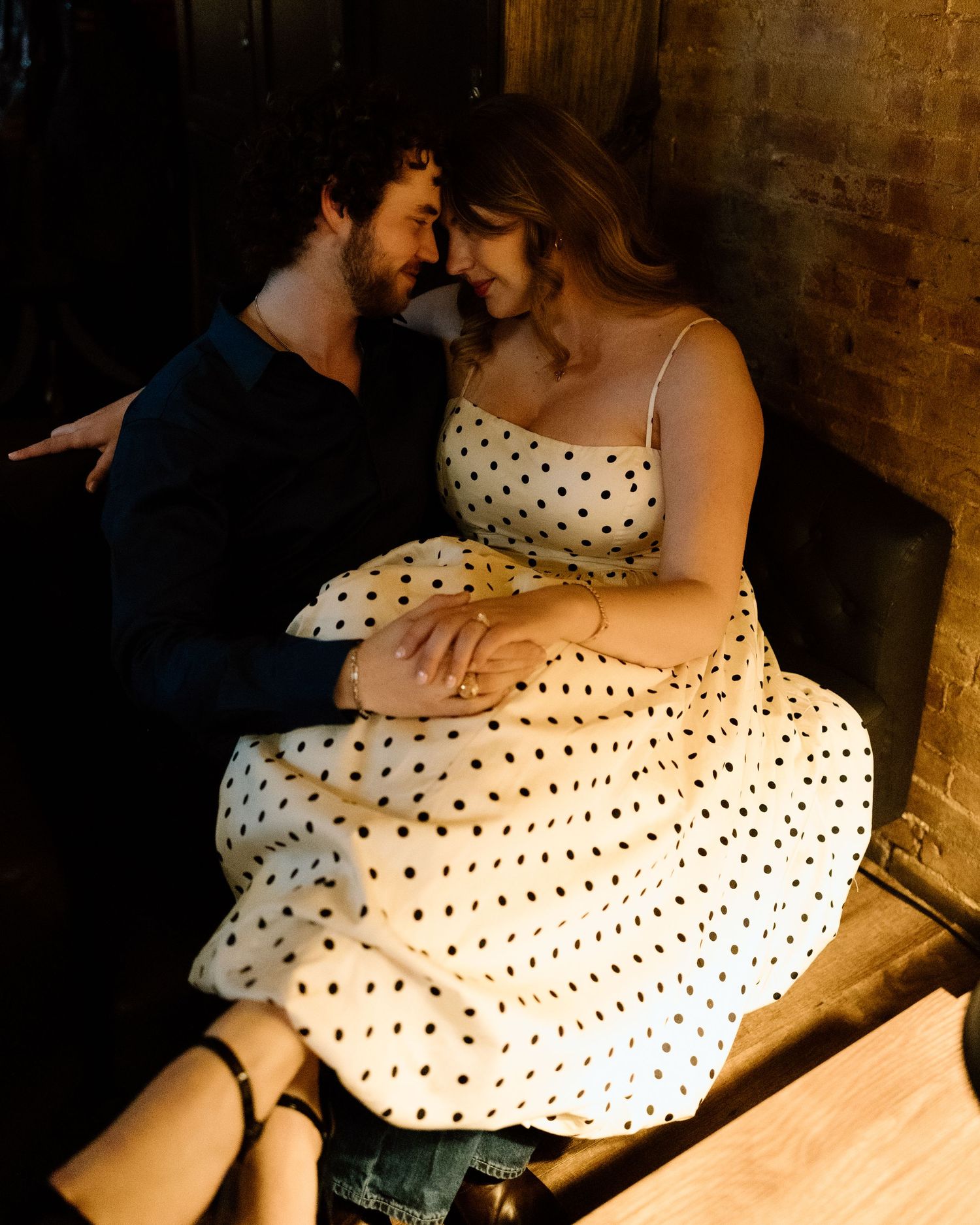 A couple in romantic embrace, woman in polka dot dress, man in navy, sharing an intimate moment in warm dim lighting.