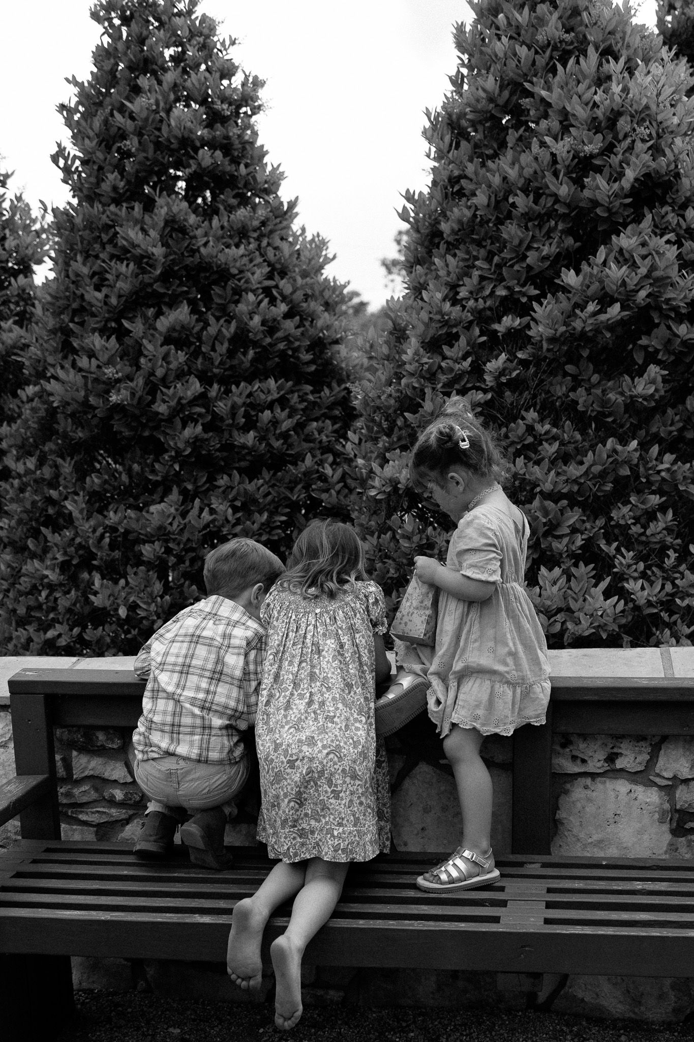 Three children sit on a bench looking over an evergreen garden in black and white.