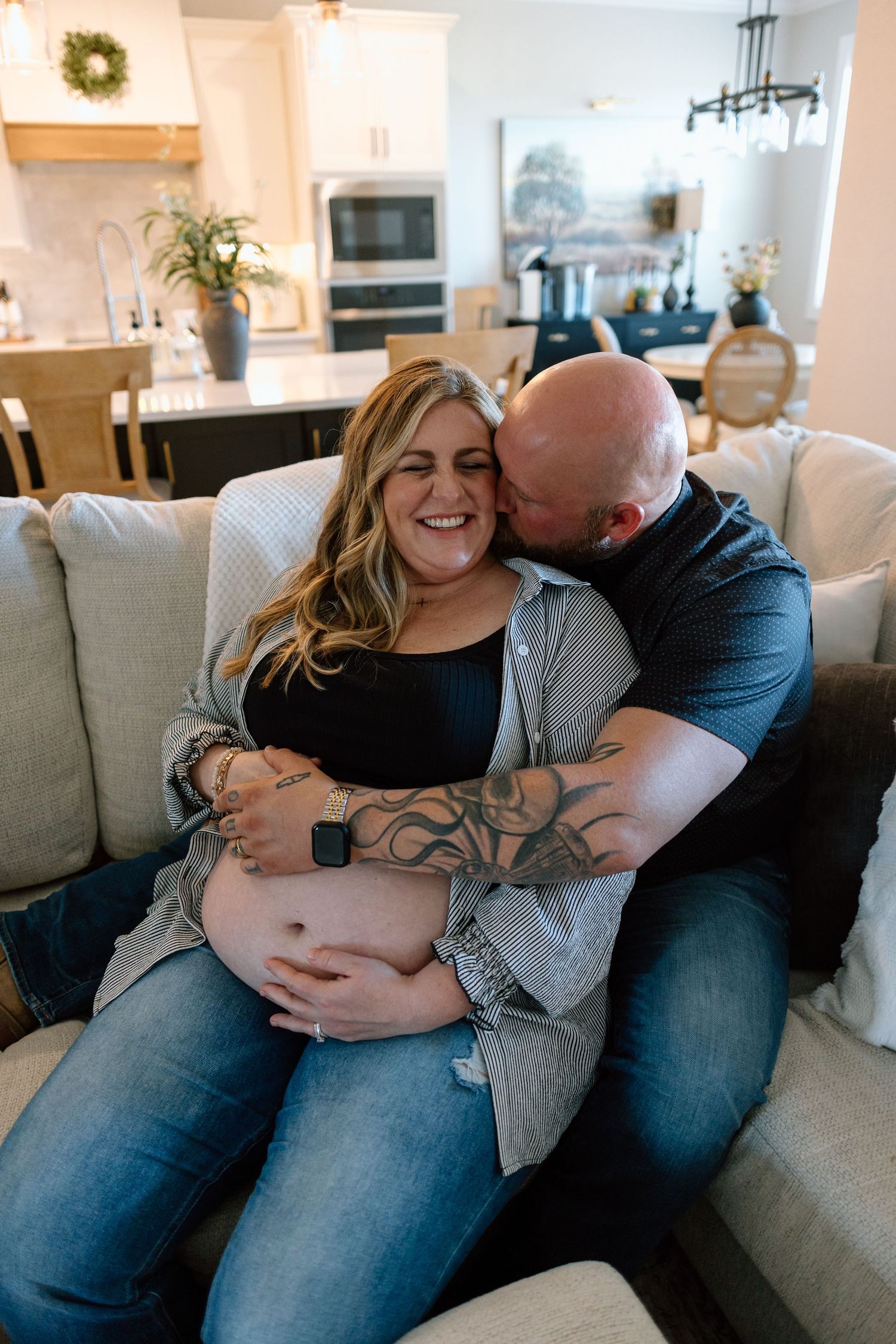 Couple cuddles on grey couch while showing off pregnancy belly in home setting.