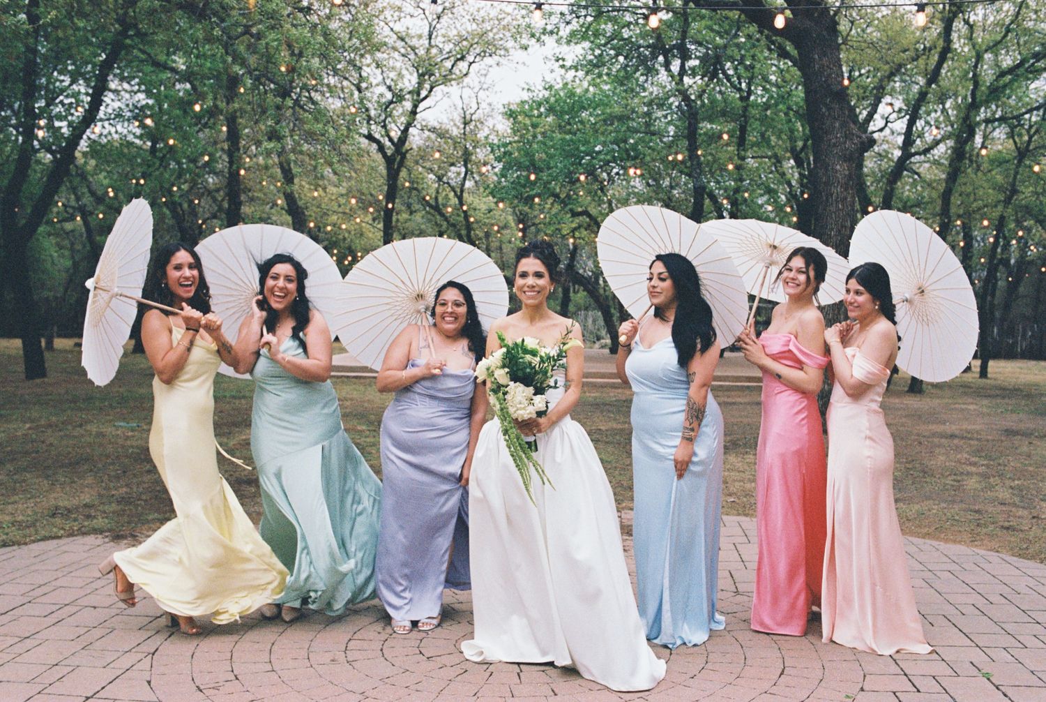 Bridal party with white parasols poses in pastel colored dresses outdoors.