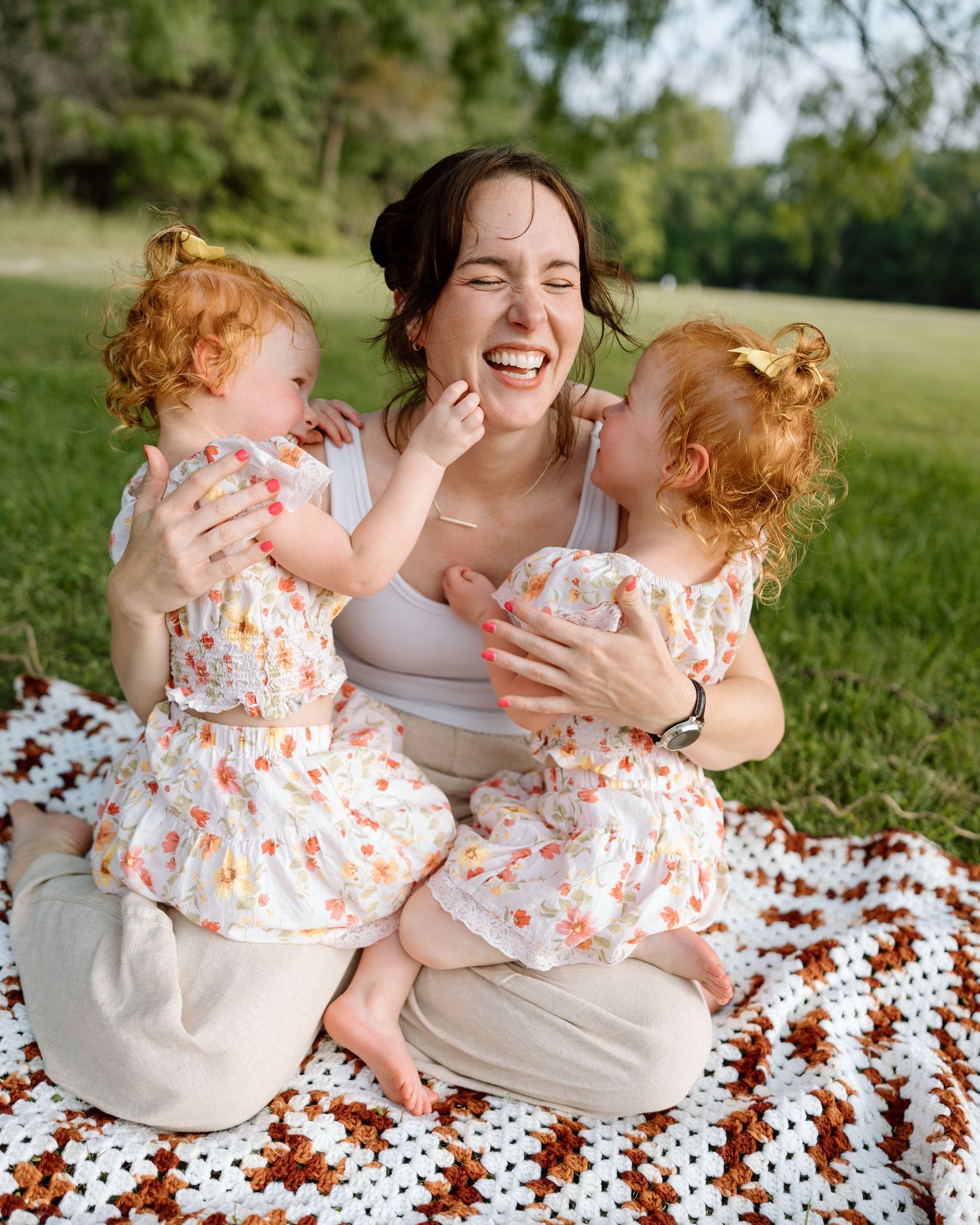 A laughing mother sits on a crochet blanket outdoors with two redhead toddler girls in matching floral dresses.