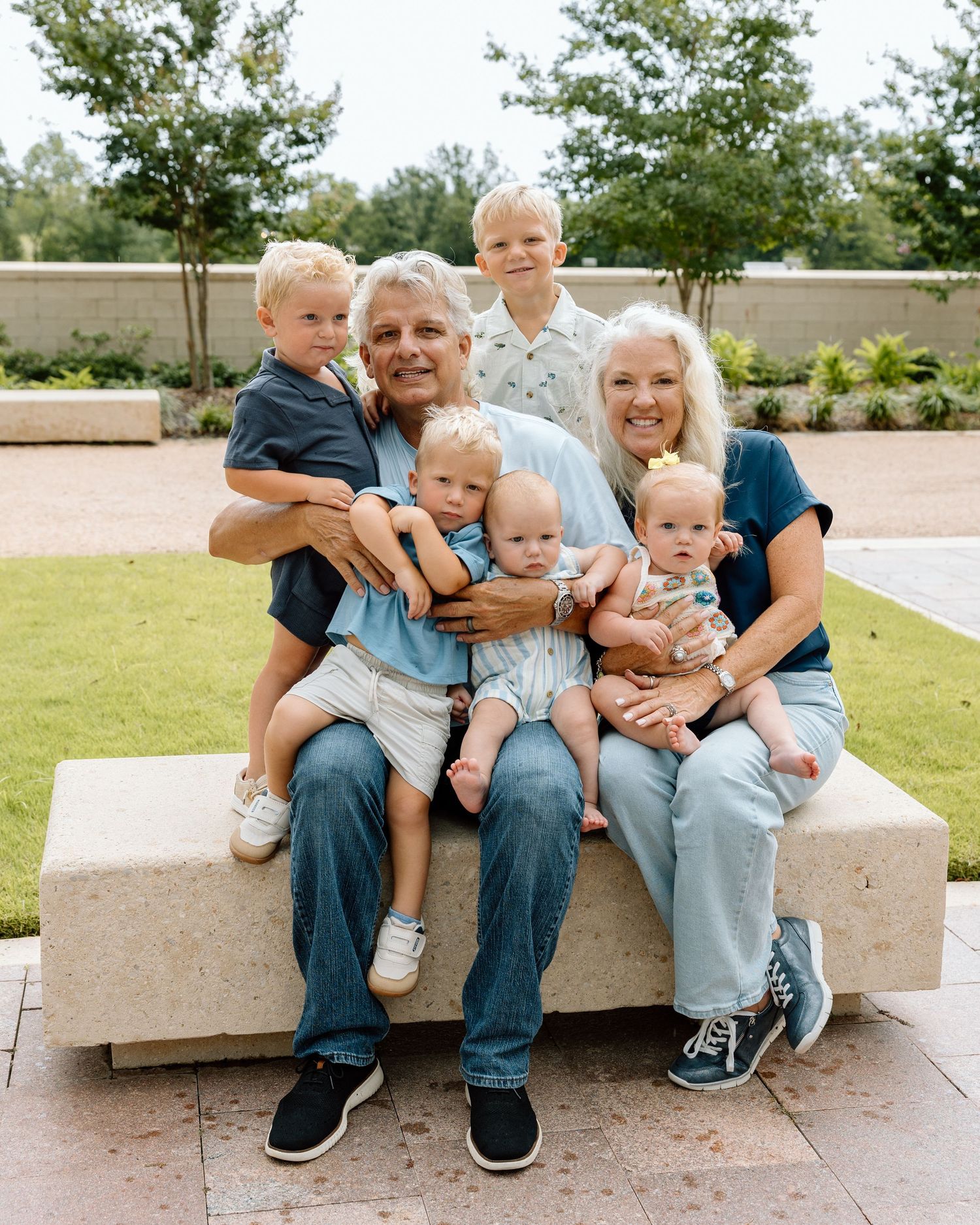 A multigenerational family poses outdoors on a stone bench, with grandparents holding three babies and two young boys standing behind them.