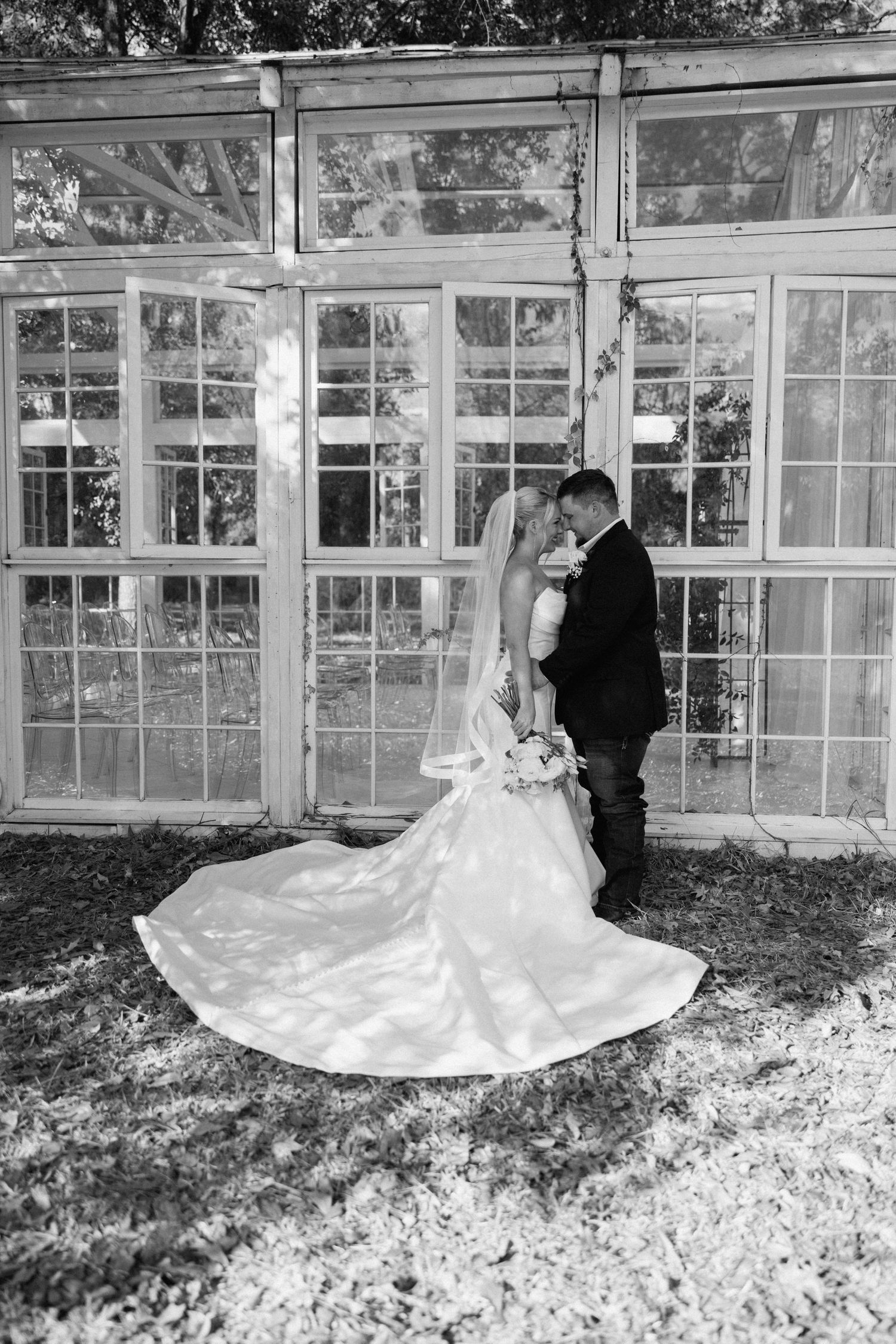 Black and white photo of bride and groom embracing before vintage greenhouse with brides long train spread on ground.
