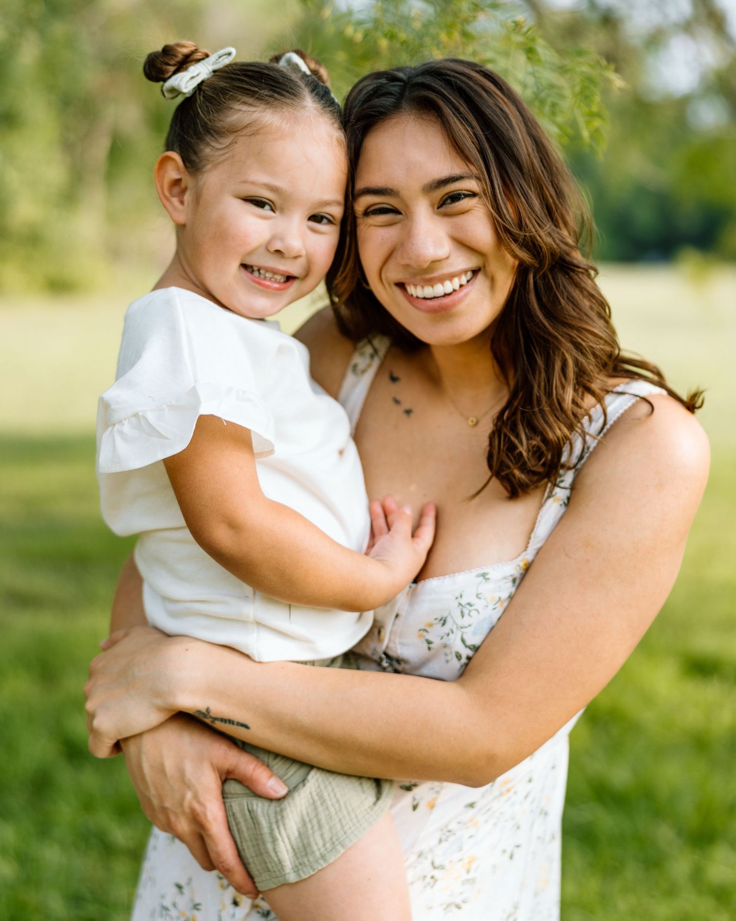 A joyful mother holds her smiling toddler daughter outdoors in a lush green park setting.