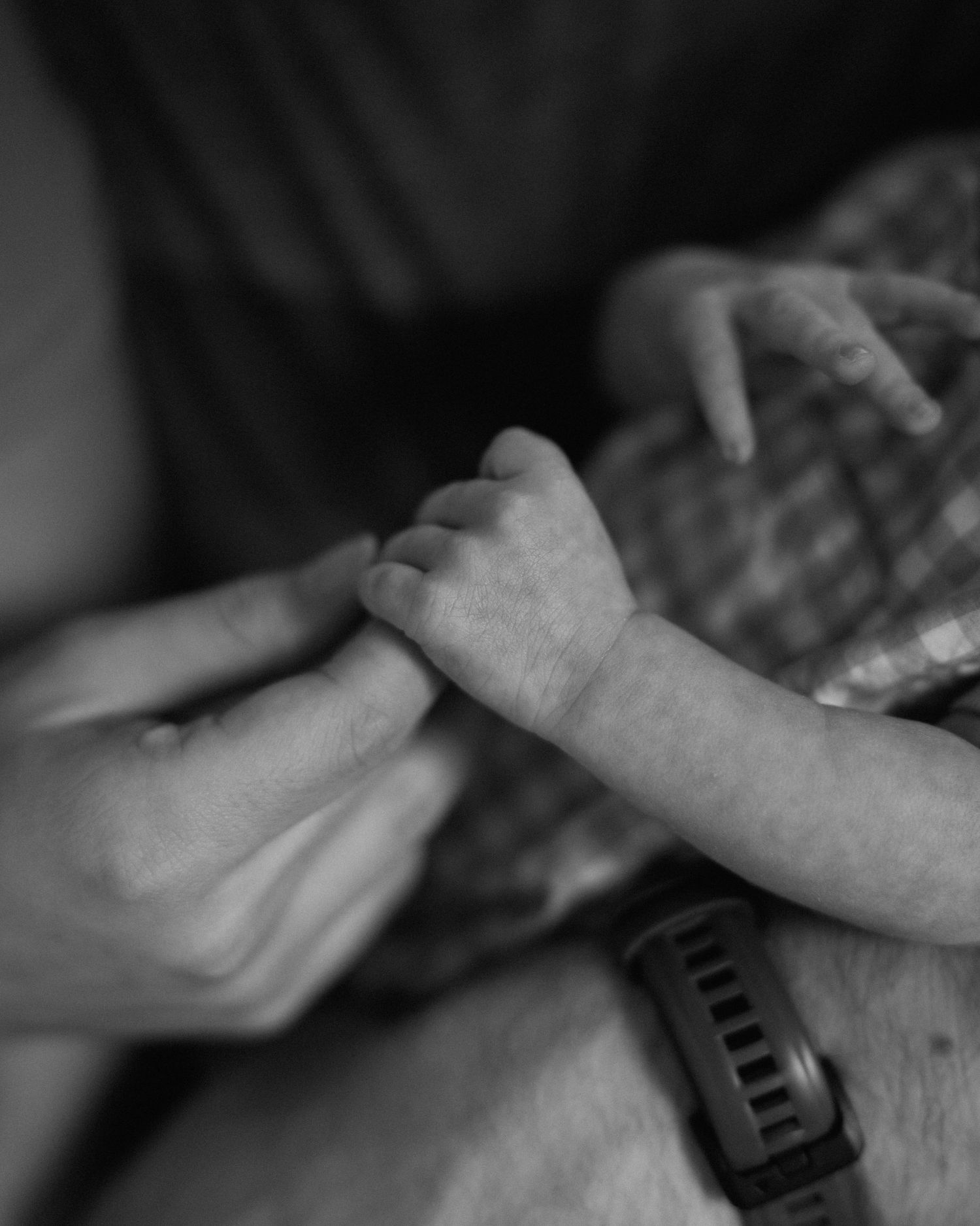 Black and white photo of an adult gently holding a newborn baby's tiny hand, wearing a smartwatch.
