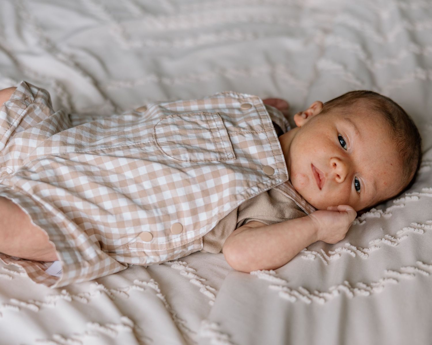 Newborn baby in plaid onesie lying on white quilted blanket, looking at camera with curious expression.