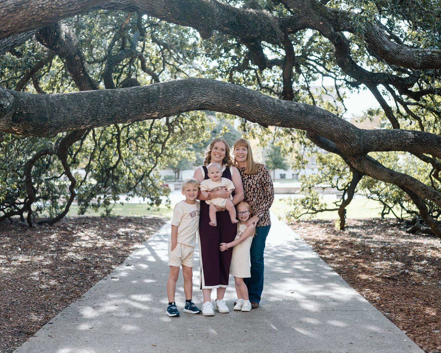 Family of five posing on a tree-lined path under a large oak tree canopy in a park setting.