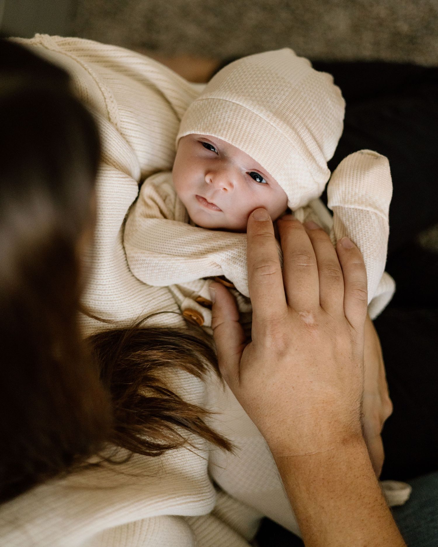 Newborn baby in cream knit outfit and hat being cradled by parents hands.