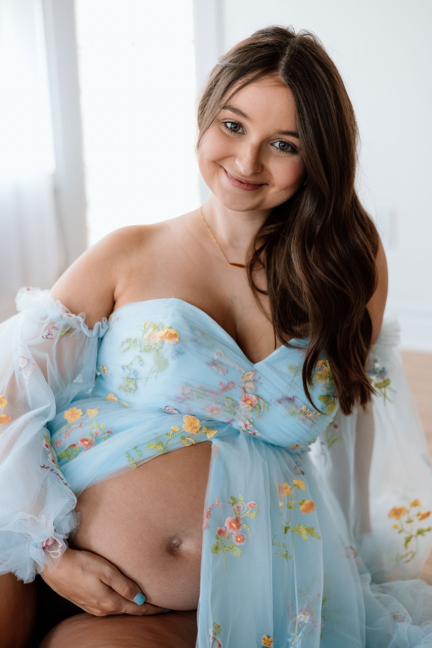 Smiling pregnant woman in a blue floral off-shoulder gown cradling her baby bump during a maternity photoshoot.