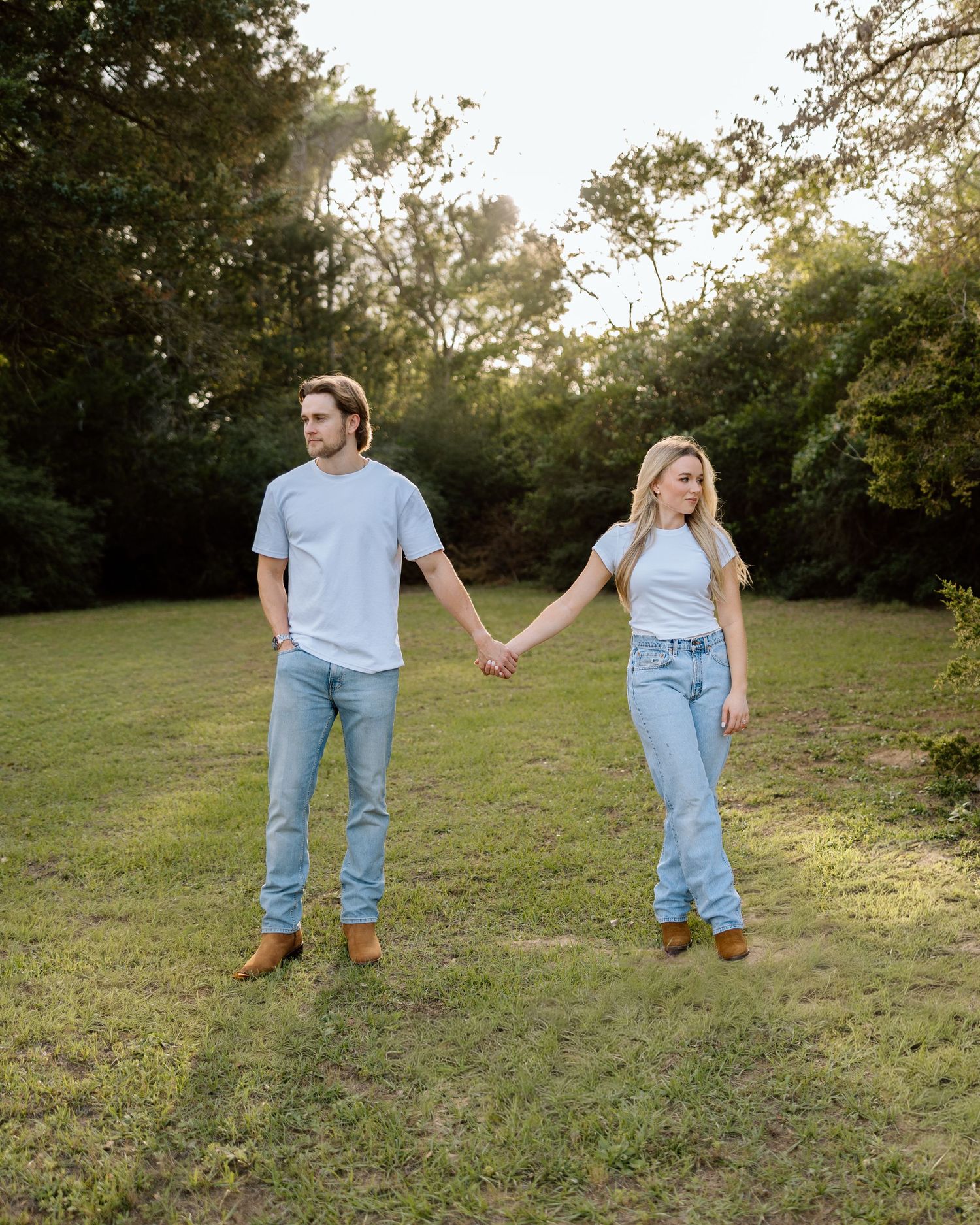 Couple holding hands in a sunlit grassy field, wearing matching white tees, light jeans, and brown boots.