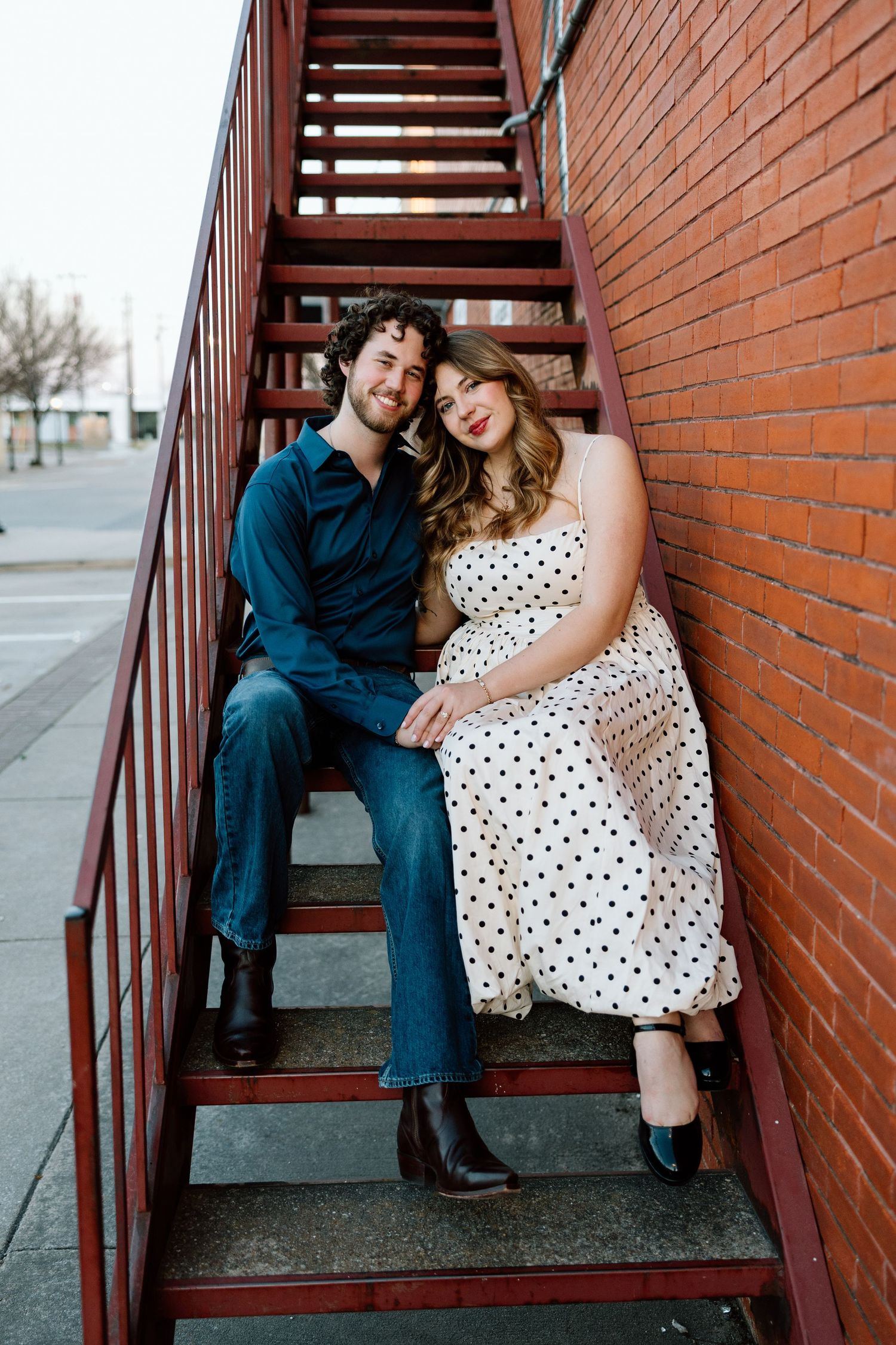 Couple sitting on red metal fire escape stairs against brick wall, woman in polka dot dress, man in teal shirt.